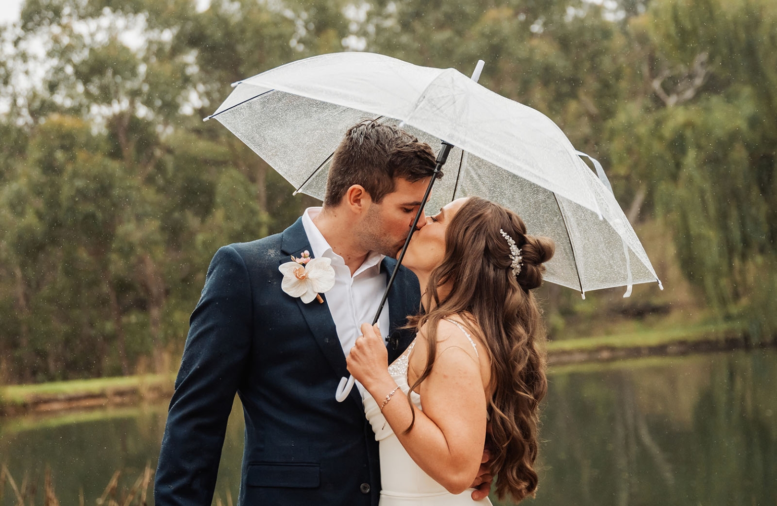 the lane vineyard wedding - caitlyn & craig 102 Wedding couple kissing under clear umbrella in rainy, lush green setting.