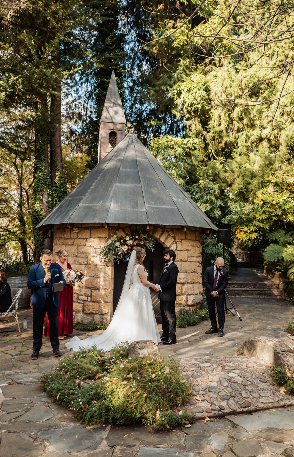 beautiful manor basket range wedding - sophie & theodore 104 Rustic outdoor wedding at stone chapel with couple, officiant, and bridesmaid amid lush greenery.