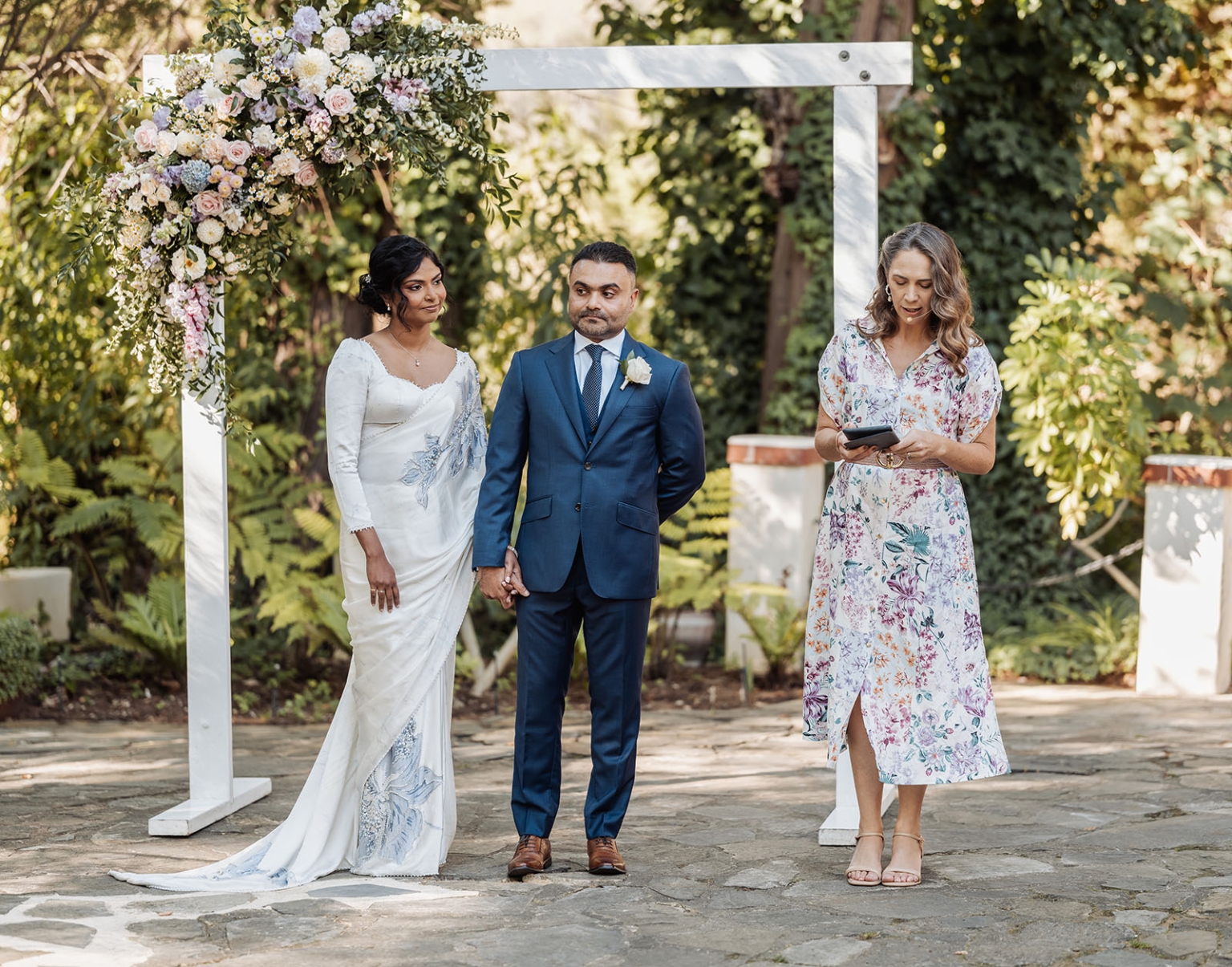 manor wedding in basket range - methmini & aravinda 136 Outdoor wedding with floral arch, couple in formal attire, lush greenery, sunlight filtering through.