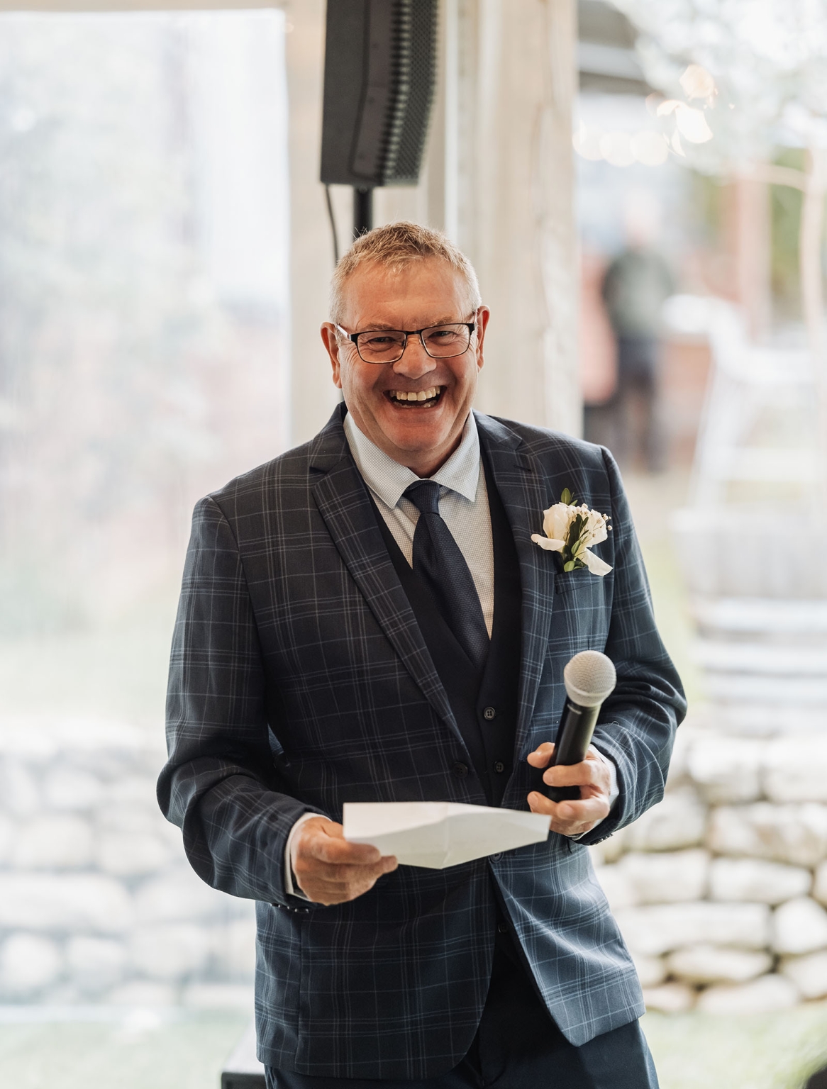 comida wedding - georgia & james 46 Older man in suit holding microphone at formal outdoor event, smiling joyfully.