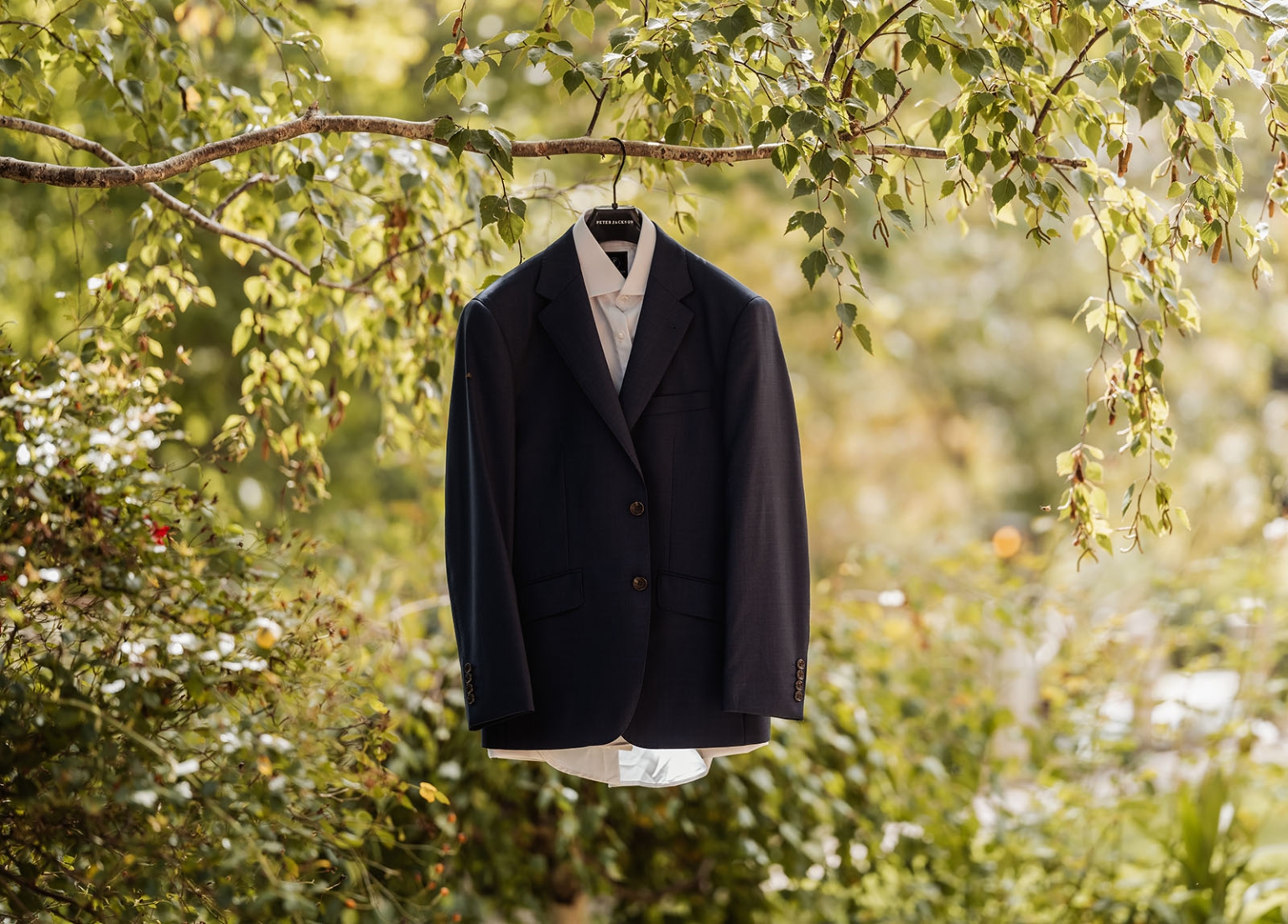 manor wedding in basket range - methmini & aravinda 94 Formal suit hanging on tree in sunny garden, contrasting nature with elegance.