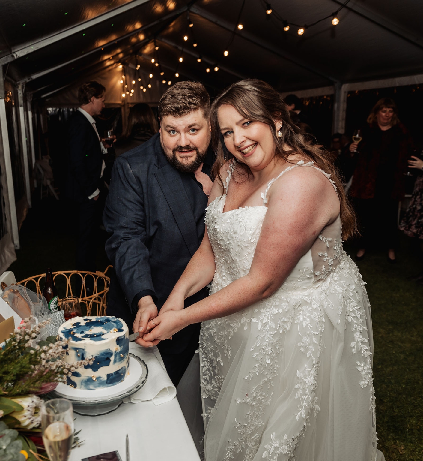 comida wedding - georgia & james 51 Bride and groom cutting elegant wedding cake under festive tent with floral decorations.