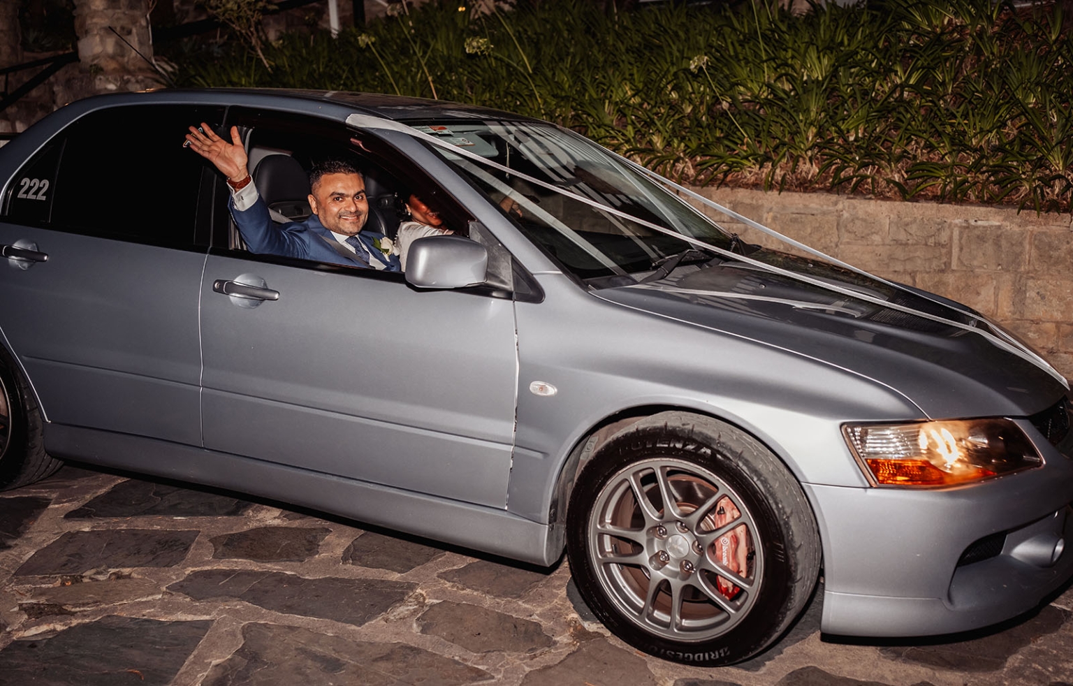 manor wedding in basket range - methmini & aravinda 177 Groom in blue suit joyfully waves from a sleek silver car, wedding celebration ambiance.