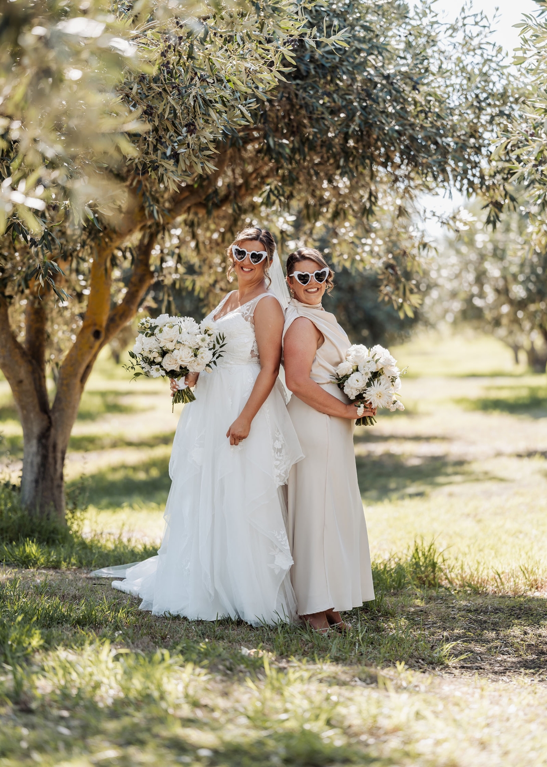 lloyd brothers wedding - dajana & tom 34 Two women at wedding, heart sunglasses, floral bouquets, garden setting, joyful celebration.
