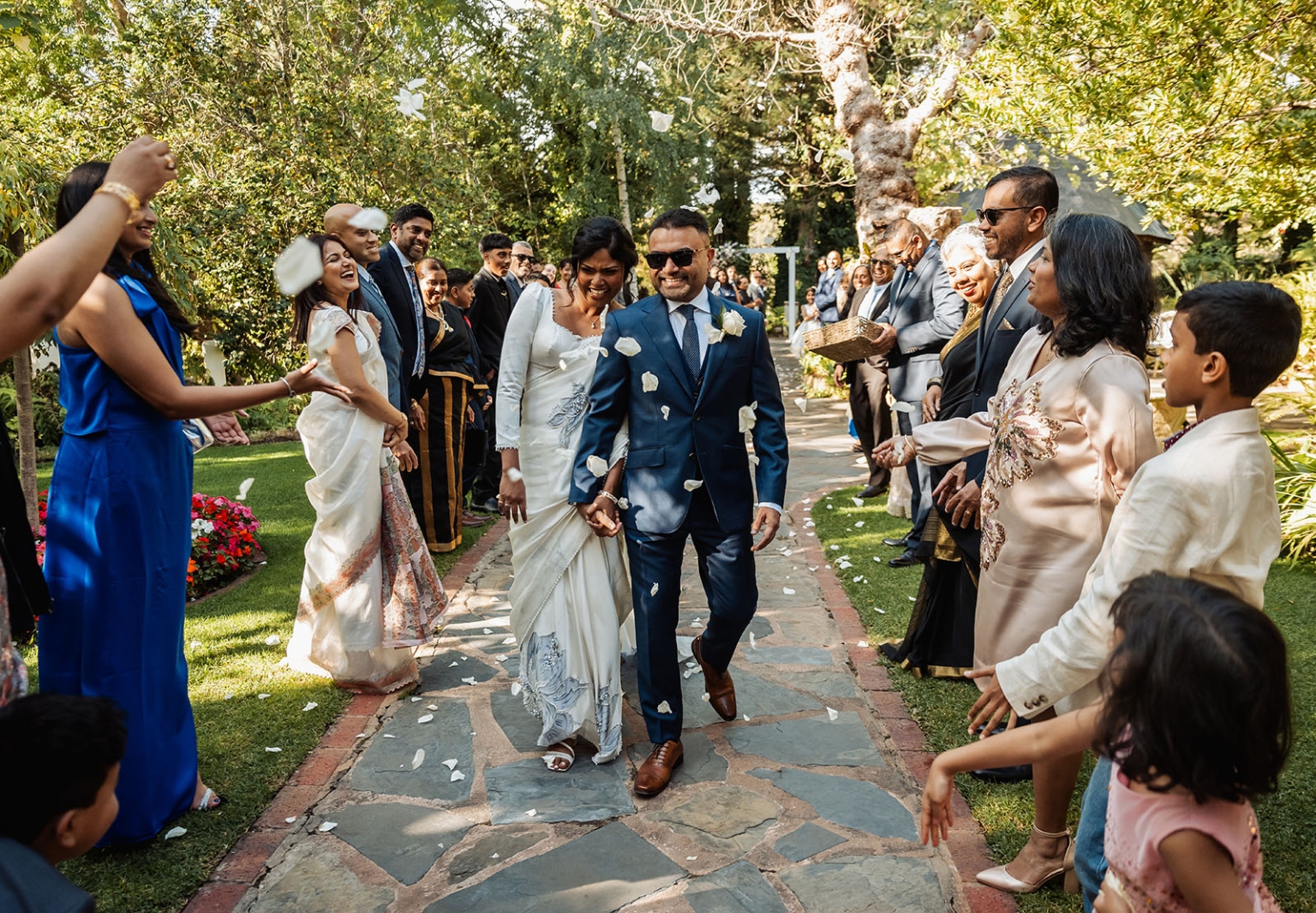 manor wedding in basket range - methmini & aravinda 143 Outdoor wedding scene with joyful newlyweds, guests, flower petals, and lush garden backdrop.