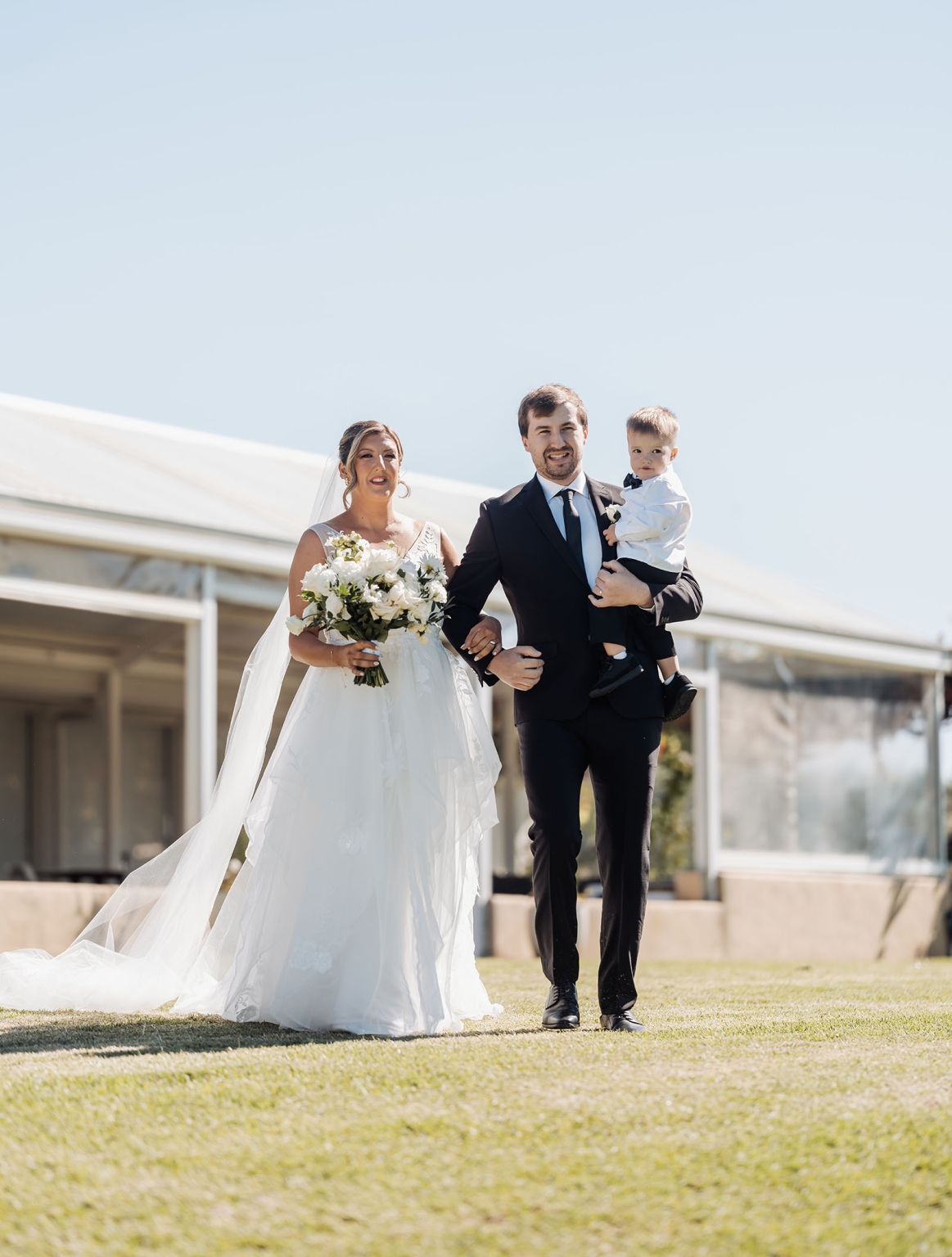 lloyd brothers wedding - dajana & tom 9 Bride and groom walking joyfully, celebrating wedding with child, elegant attire, sunny day.