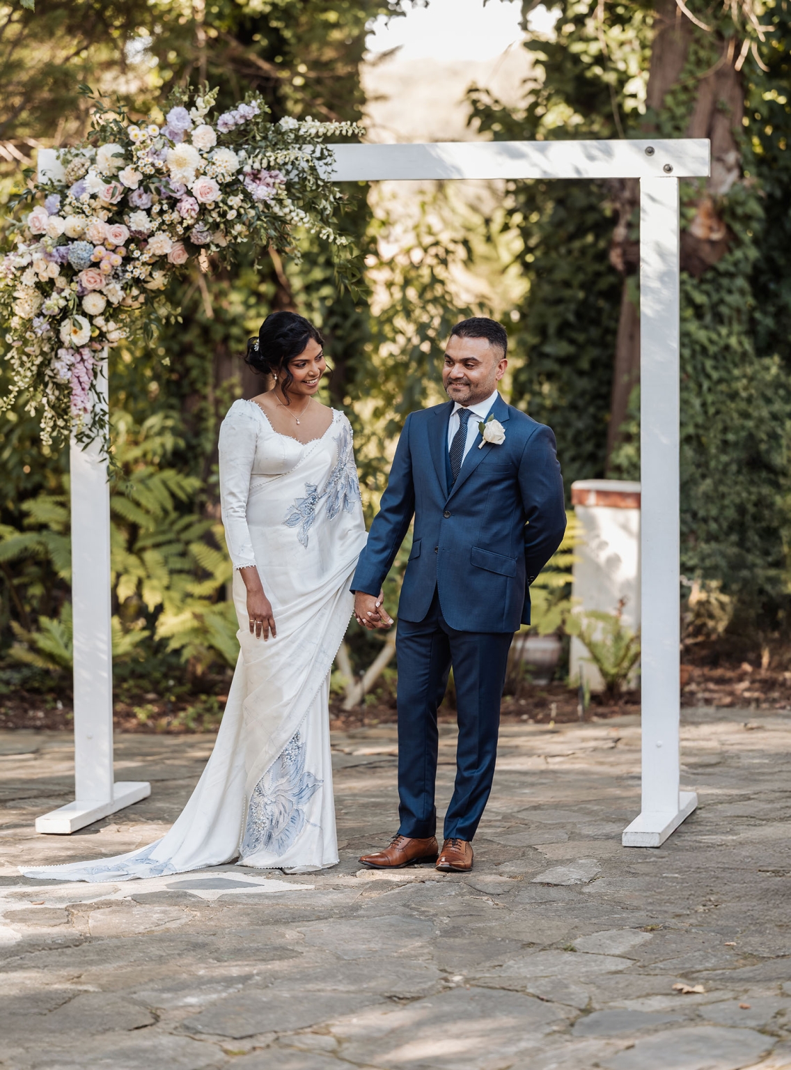 manor wedding in basket range - methmini & aravinda 137 Couple under floral arch in garden, wearing saree and suit, celebrating wedding.