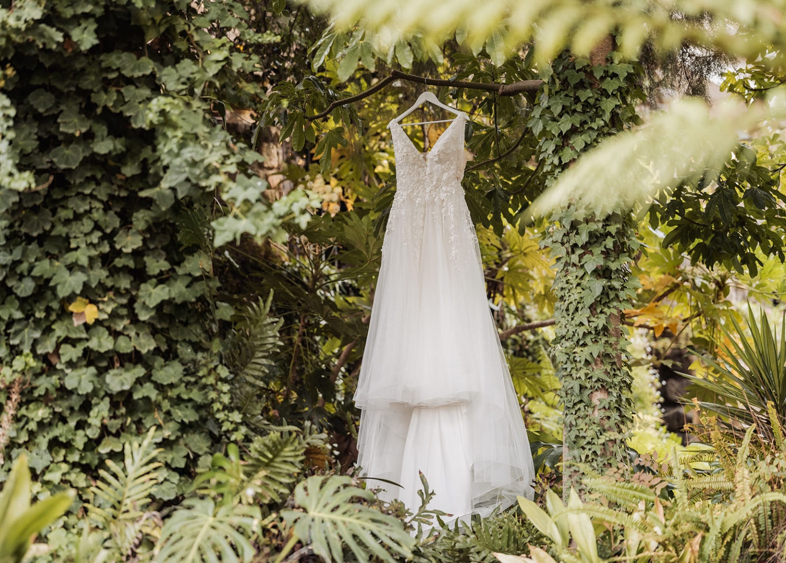beautiful manor basket range wedding - sophie & theodore 89 Elegant white wedding dress hanging in lush garden with green foliage.