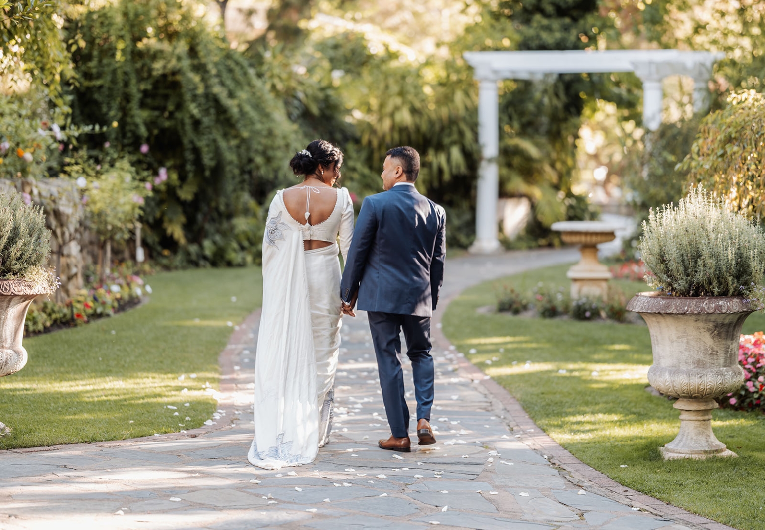 manor wedding in basket range - methmini & aravinda 147 Bride and groom walking hand-in-hand in a lush garden pathway during wedding.