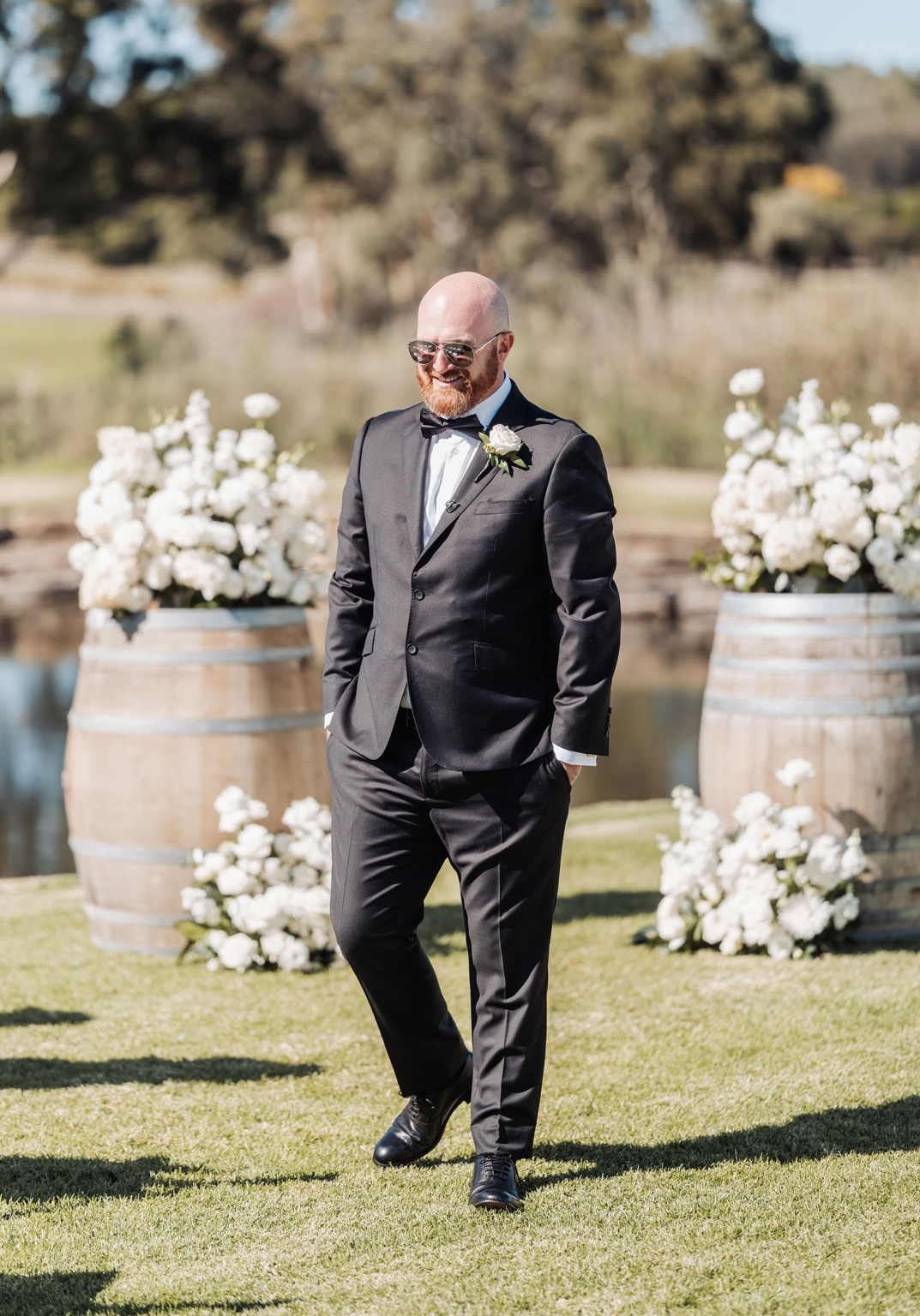 lloyd brothers wedding - dajana & tom 5 Man in black tuxedo with white rose on green lawn at outdoor wedding.