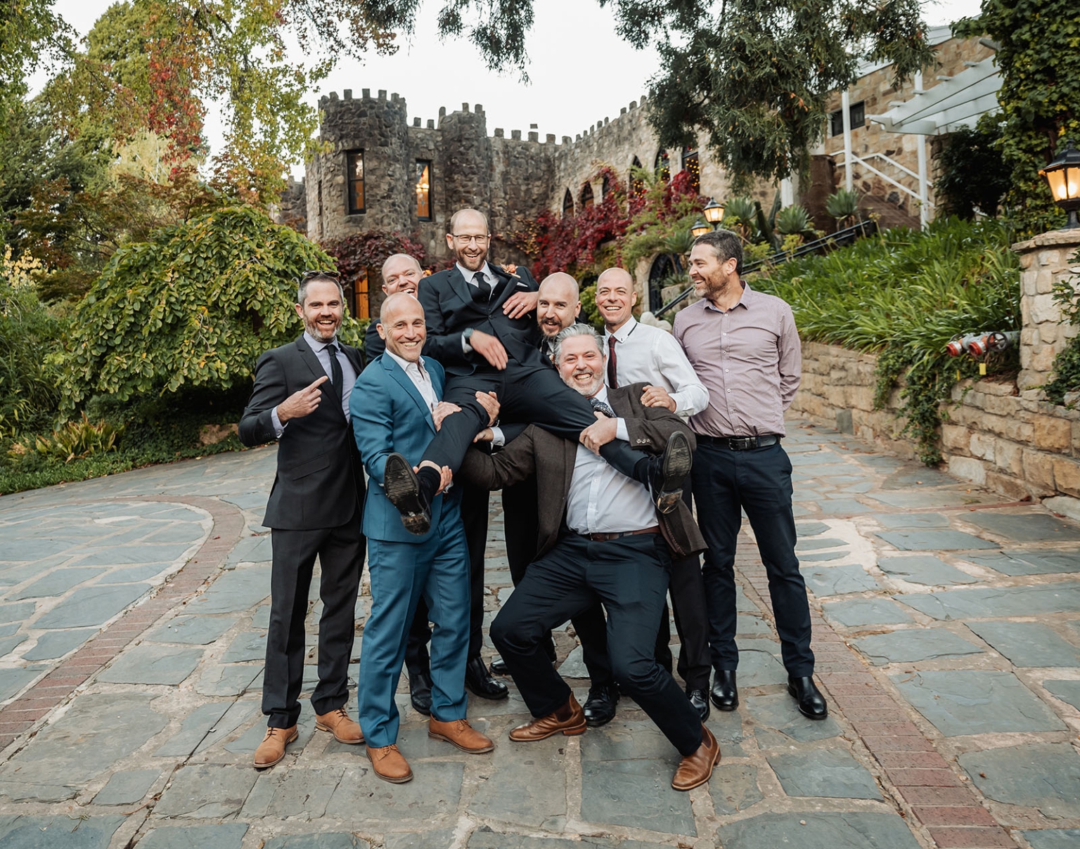 adelaide hills wedding at the manor - jo & dan 46 Groomsmen lift groom in castle courtyard during joyful wedding celebration.