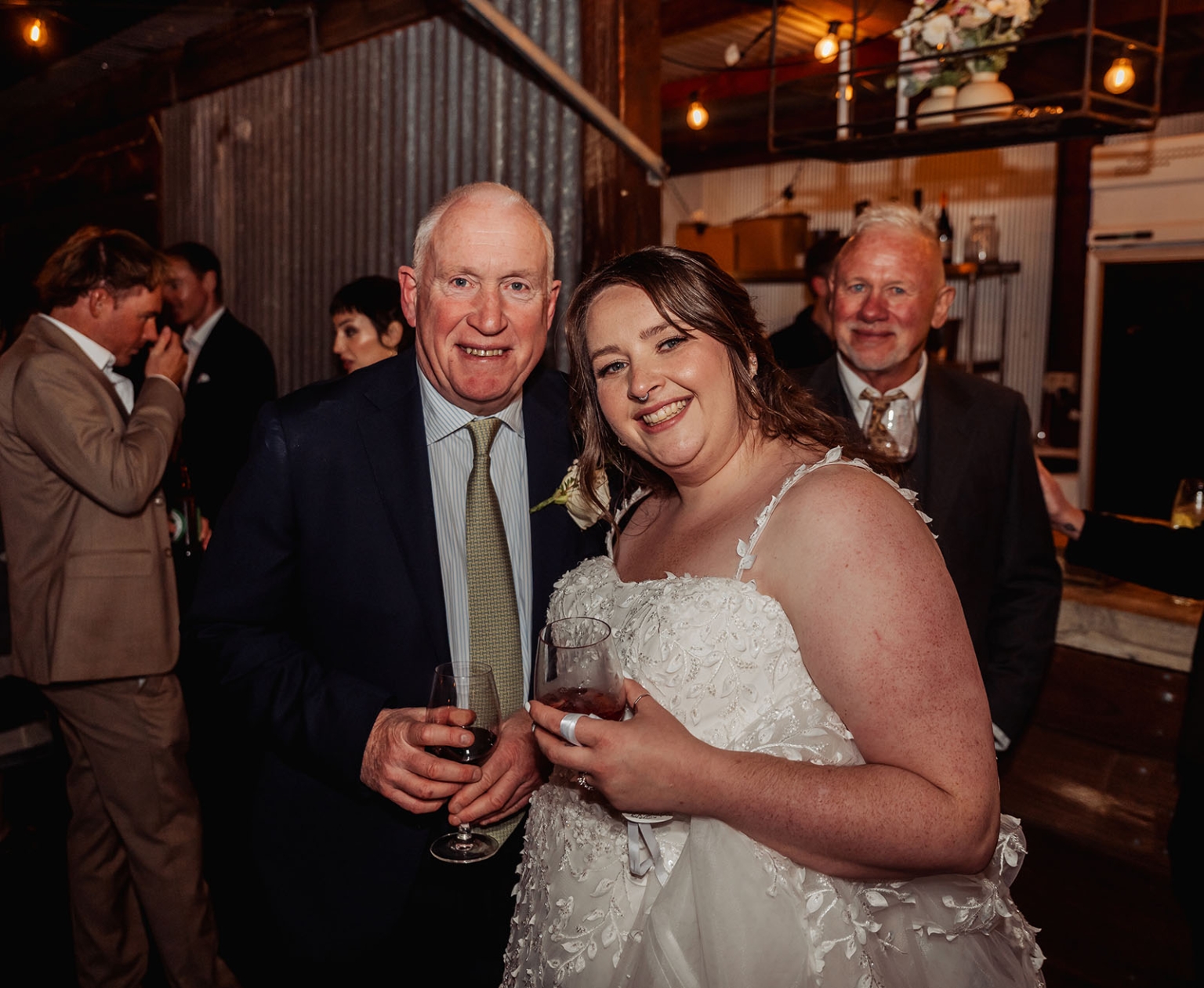 comida wedding - georgia & james 50 Smiling couple in formal attire at rustic wedding celebration with guests and floral decor.