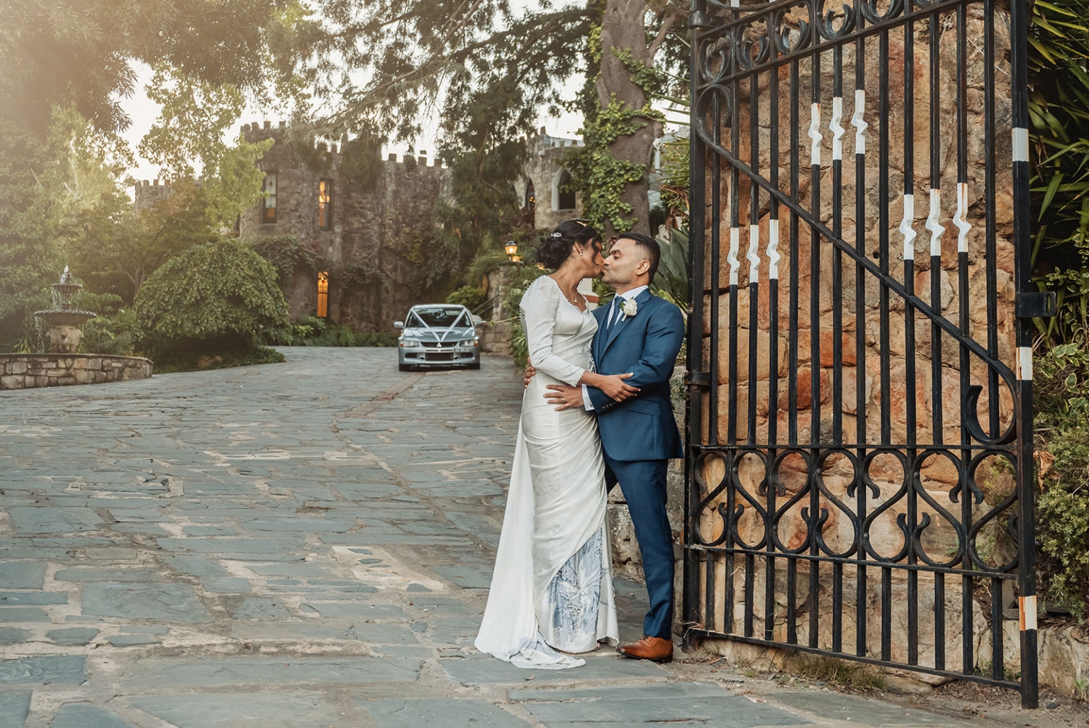 manor wedding in basket range - methmini & aravinda 160 Wedding couple embracing by castle gate with elegant attire, cobblestone path, and warm sunset.