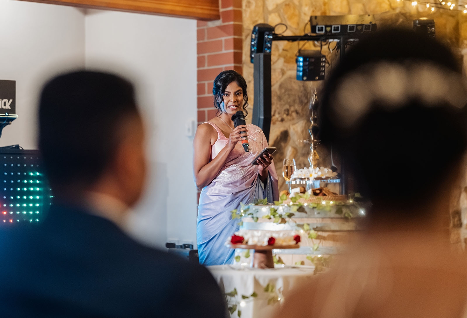 manor wedding in basket range - methmini & aravinda 166 Woman delivering wedding speech in embroidered dress, rustic venue, desserts on table, warm ambiance.