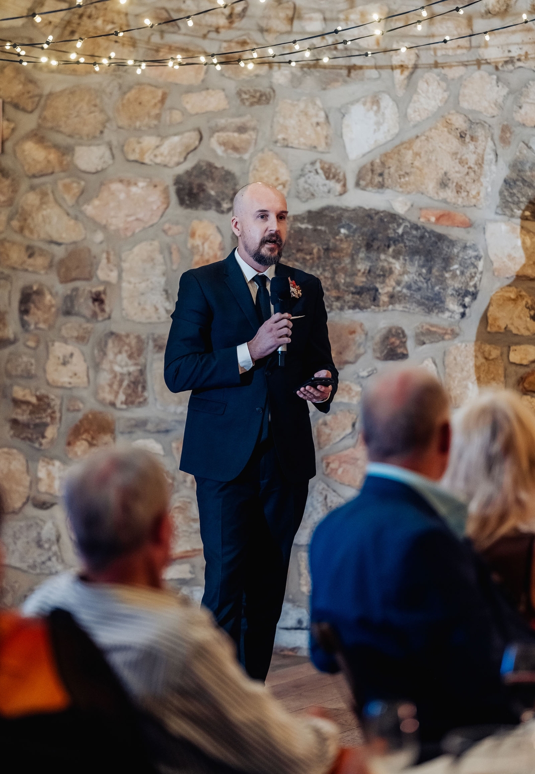 adelaide hills wedding at the manor - jo & dan 64 Man gives wedding speech before rustic stone wall with string lights.
