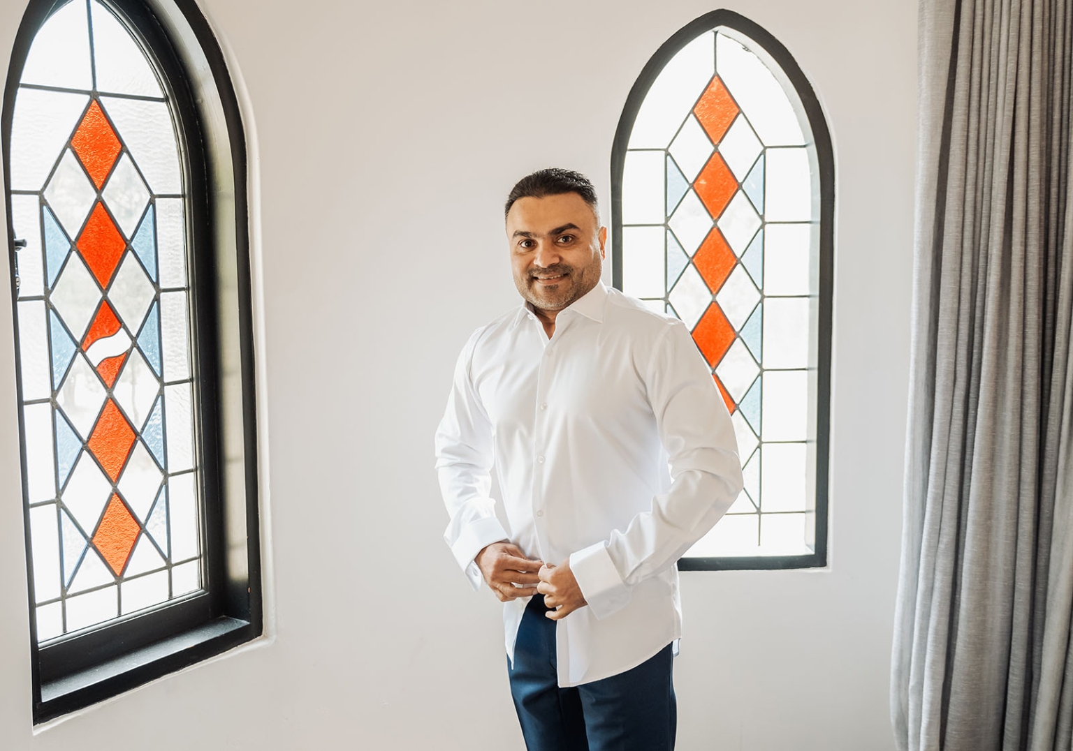 manor wedding in basket range - methmini & aravinda 96 Elegant man in formal attire by stained-glass windows in stylish, softly lit room.