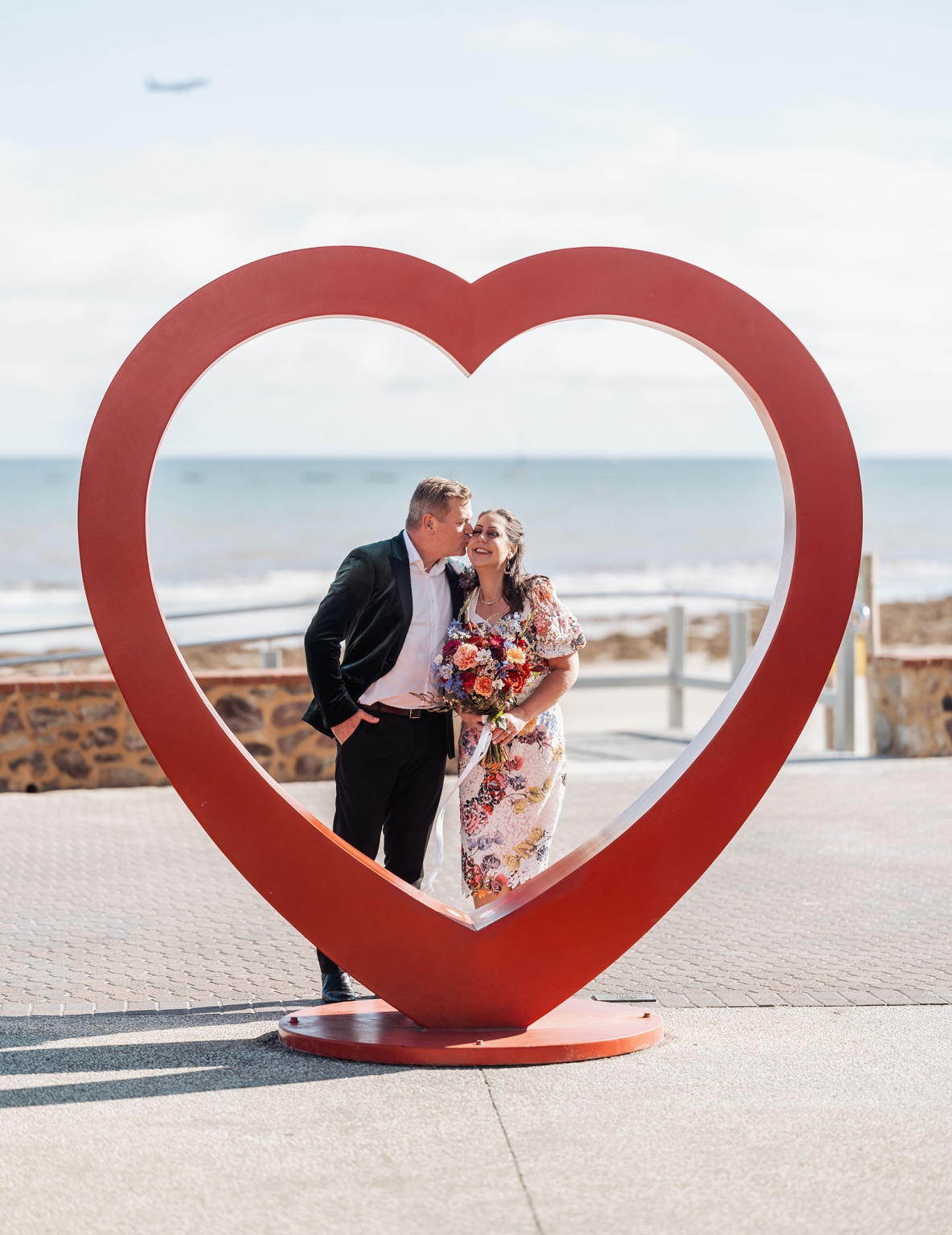sa marriage office wedding – melissa & kyran 225 Wedding couple kissing inside red heart sculpture by the ocean with colorful bouquet.