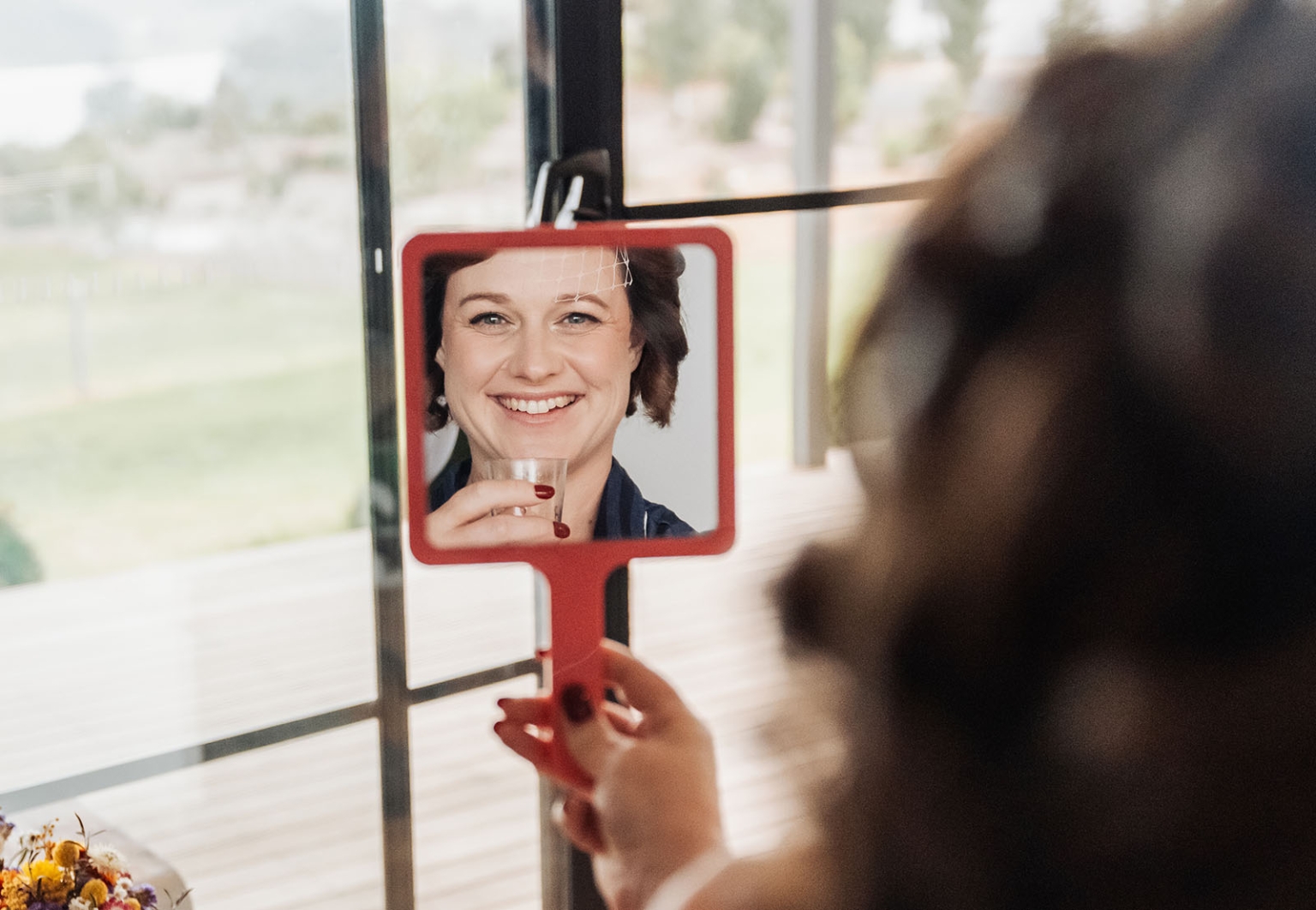 tiny house on the prairie wedding - lauren & bek 5 Bride smiling in red handheld mirror during wedding preparation.