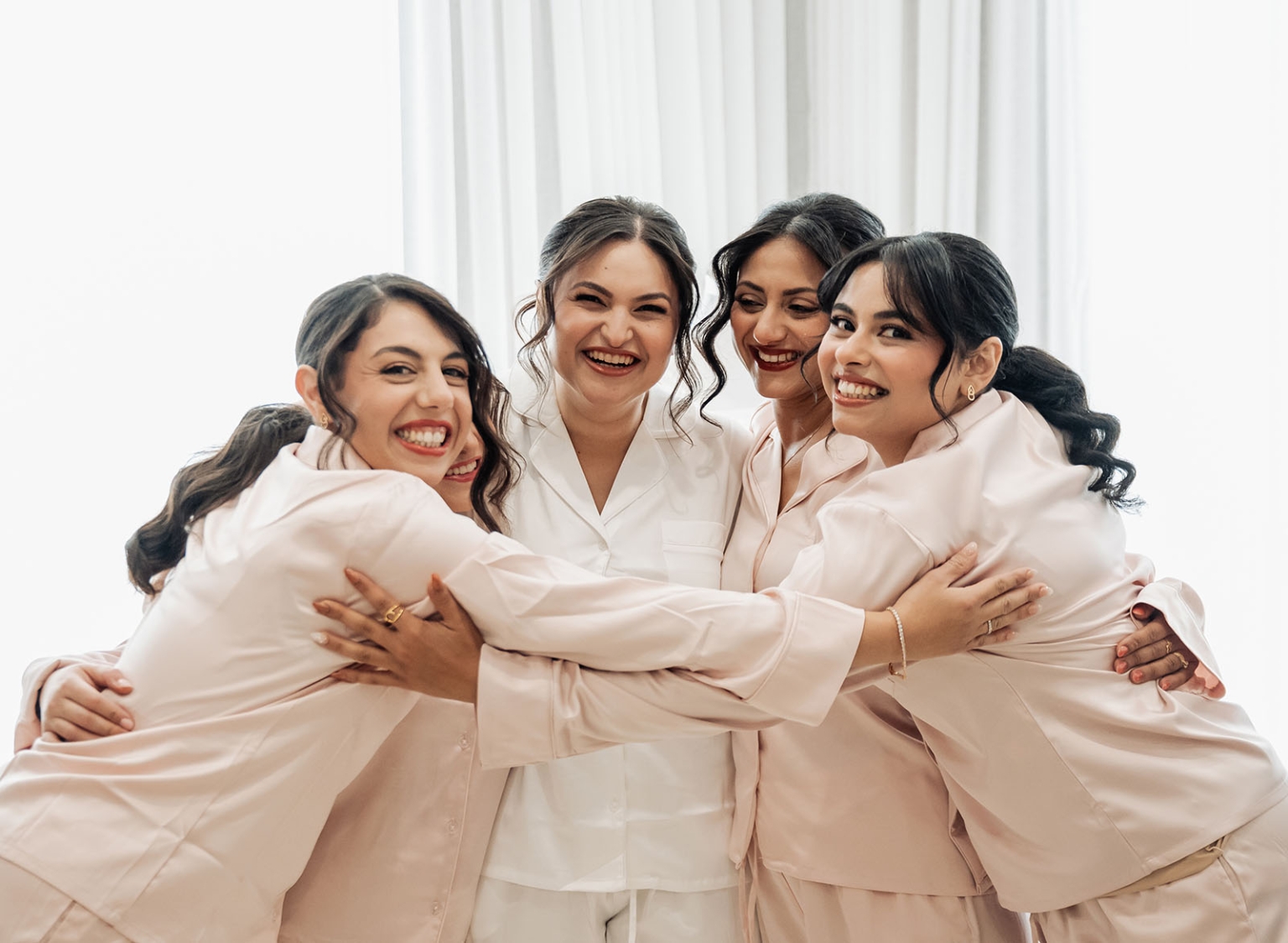 the night before the wedding: how brides can wake up with camera-ready hair 104 Bride and bridesmaids in satin pajamas sharing joyful wedding morning group hug.