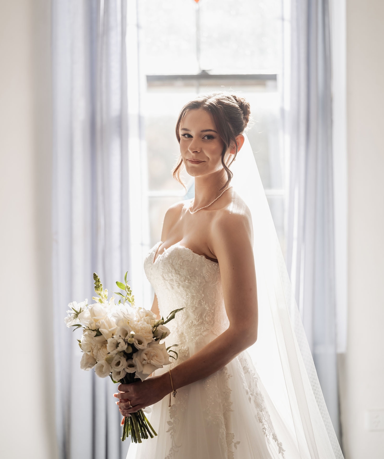 the manor wedding - crystal & mackenzie 21 Bride in lace wedding gown holding white bouquet by sunlit window.