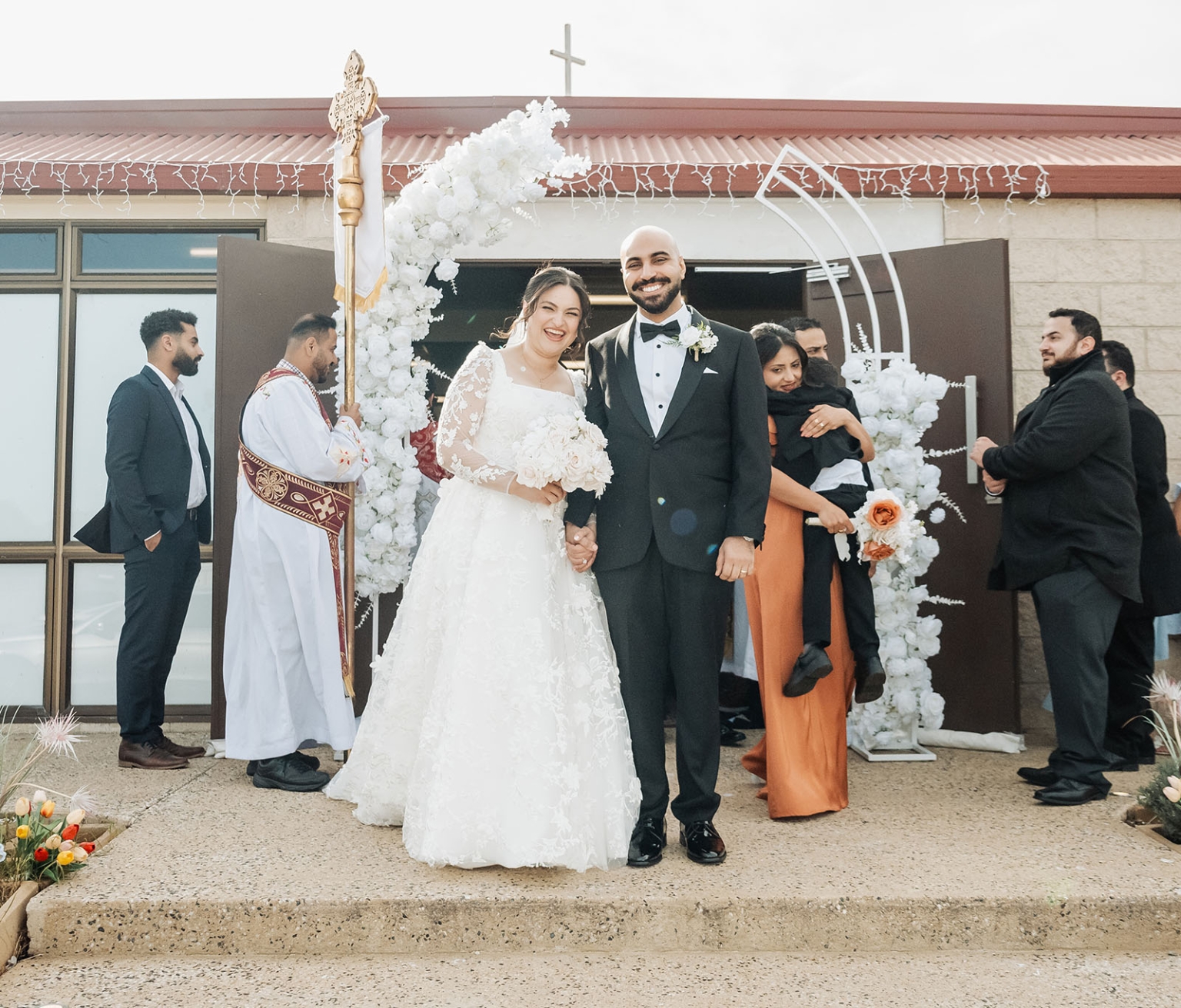 st george & st shenouda coptic church wedding - carmina & bill 53 Newlyweds smiling outside church after wedding ceremony under white floral arch.