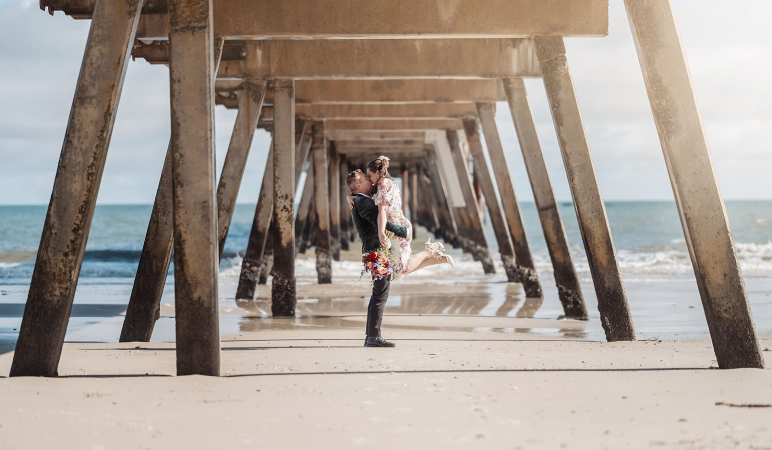sa marriage office wedding – melissa & kyran 224 Wedding couple kissing under beach pier with bouquet and ocean waves.