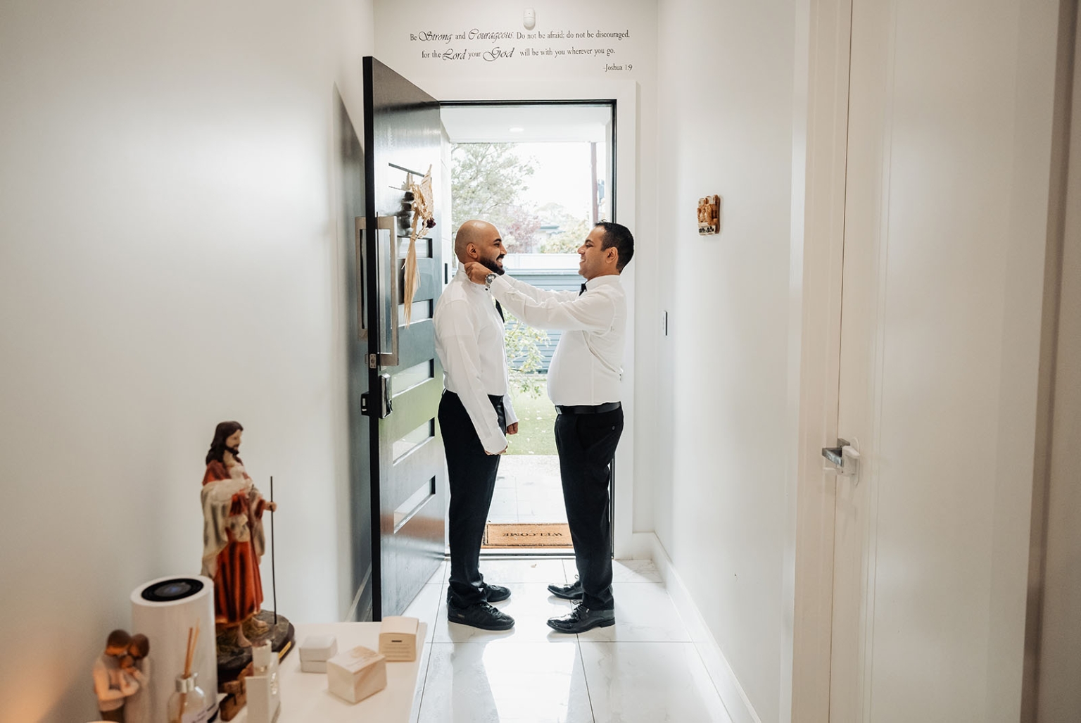 st george & st shenouda coptic church wedding - carmina & bill 5 Wedding groom collar adjustment in bright modern entryway with Joshua 1:9 wall quote.