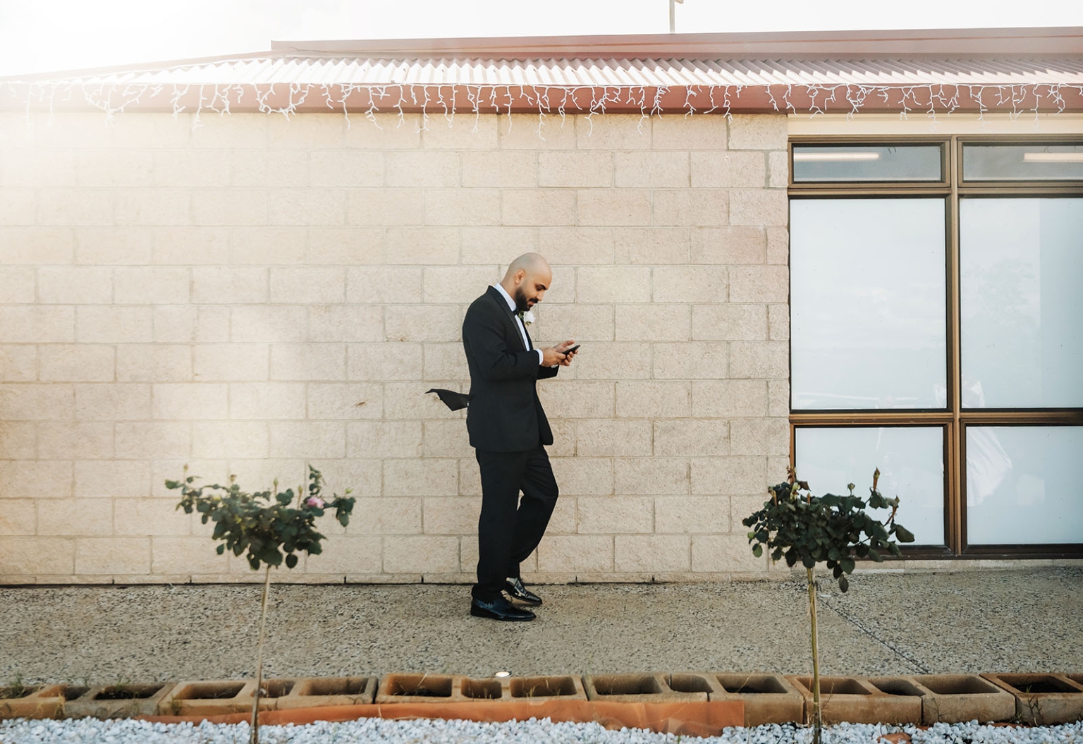 st george & st shenouda coptic church wedding - carmina & bill 33 Formal groom checking smartphone beside brick wall with wedding string lights.
