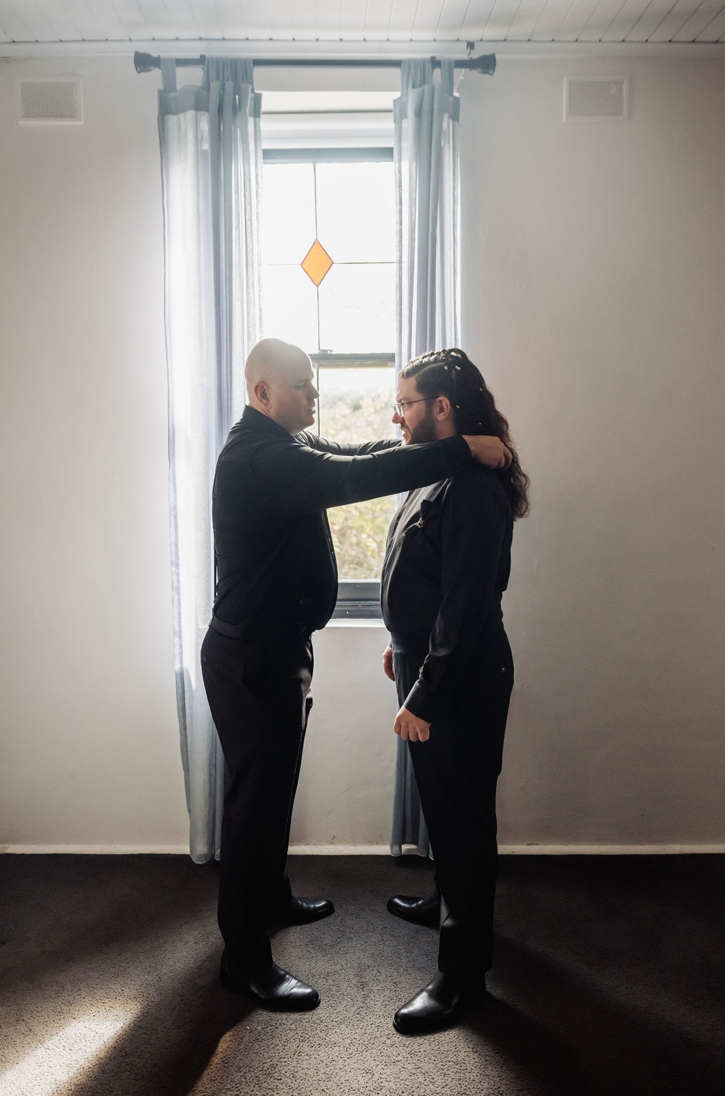 basket range wedding at the manor - leah & wes 21 Two men in black formalwear preparing by a bright stained-glass window.