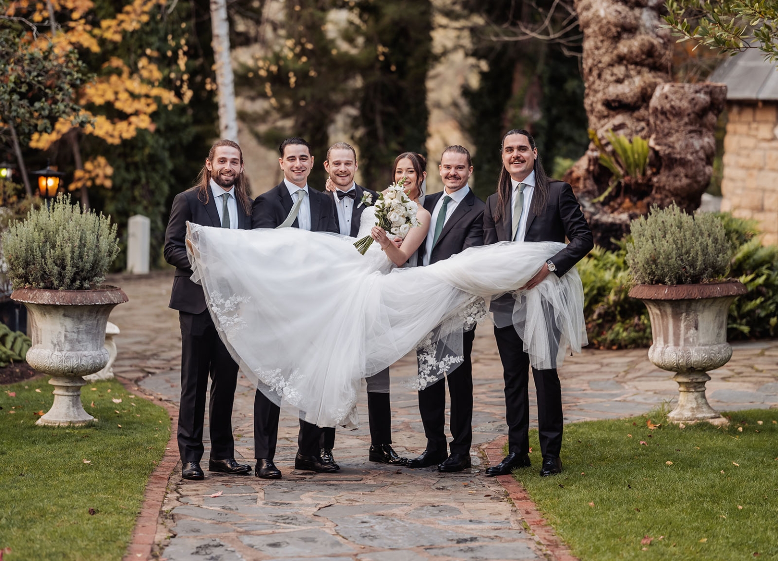 the manor wedding - crystal & mackenzie 71 Garden wedding bridal party lifts smiling bride in white gown on stone pathway.