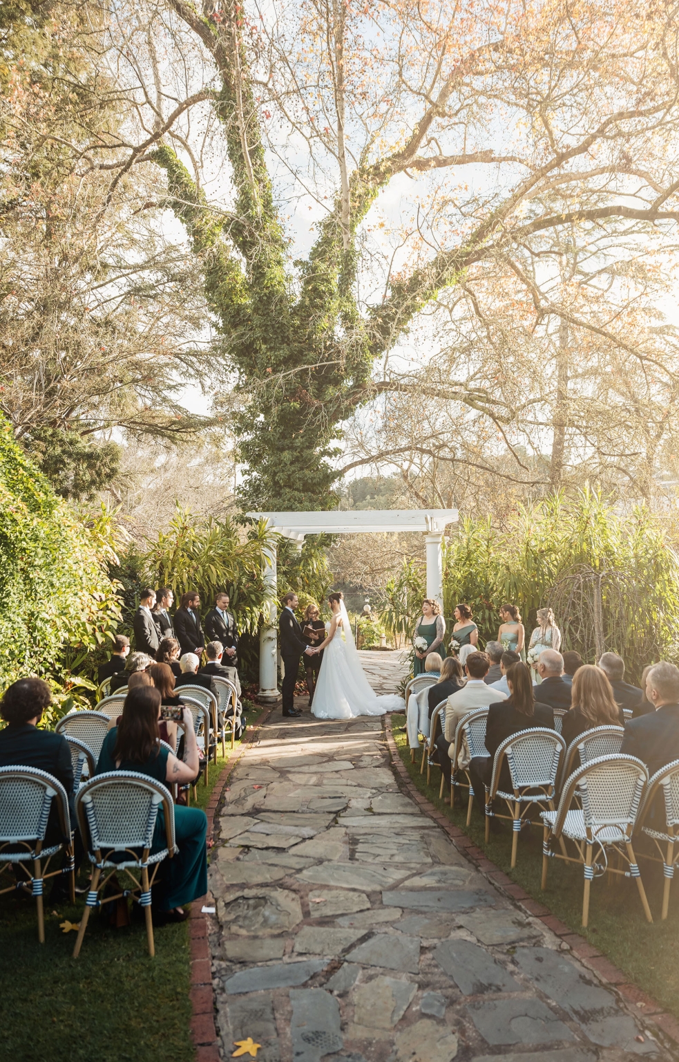 the manor wedding - crystal & mackenzie 51 Garden wedding ceremony under sprawling tree with bride, groom, guests, and bridal party.
