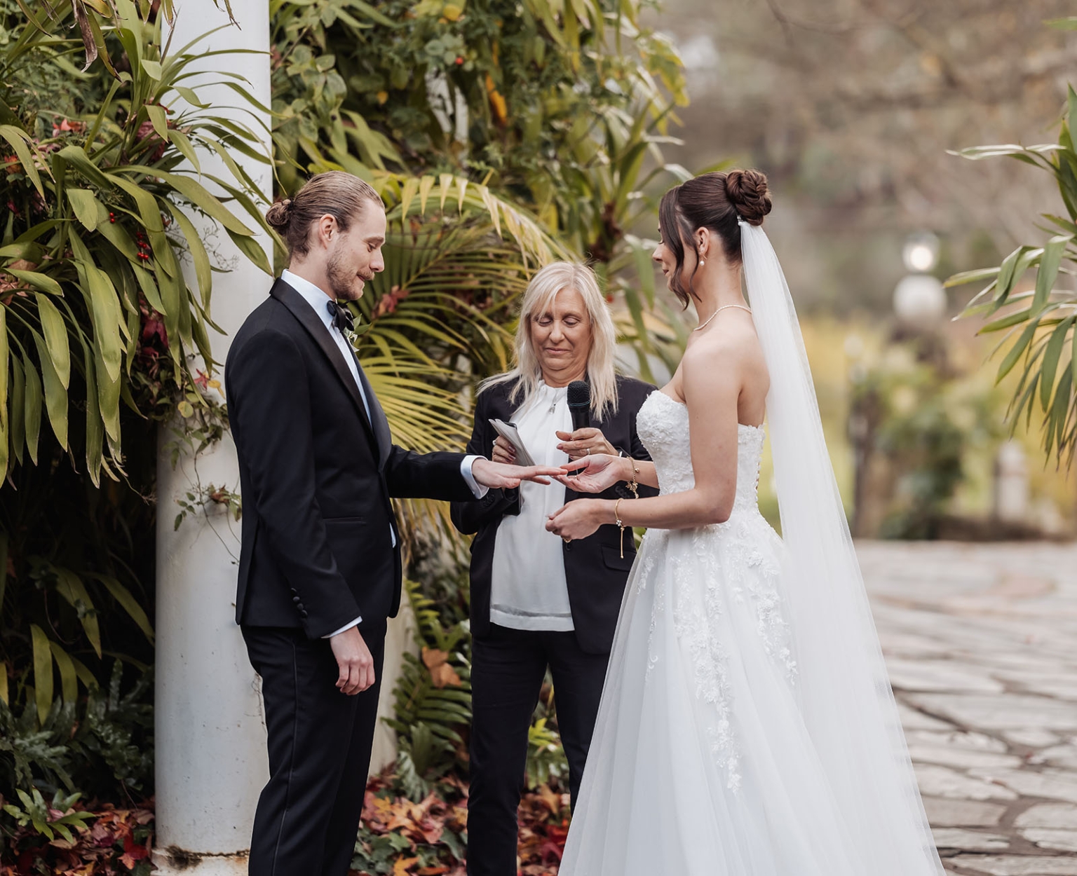 the manor wedding - crystal & mackenzie 56 Garden wedding ring exchange with bride, groom, and officiant in tropical greenery.