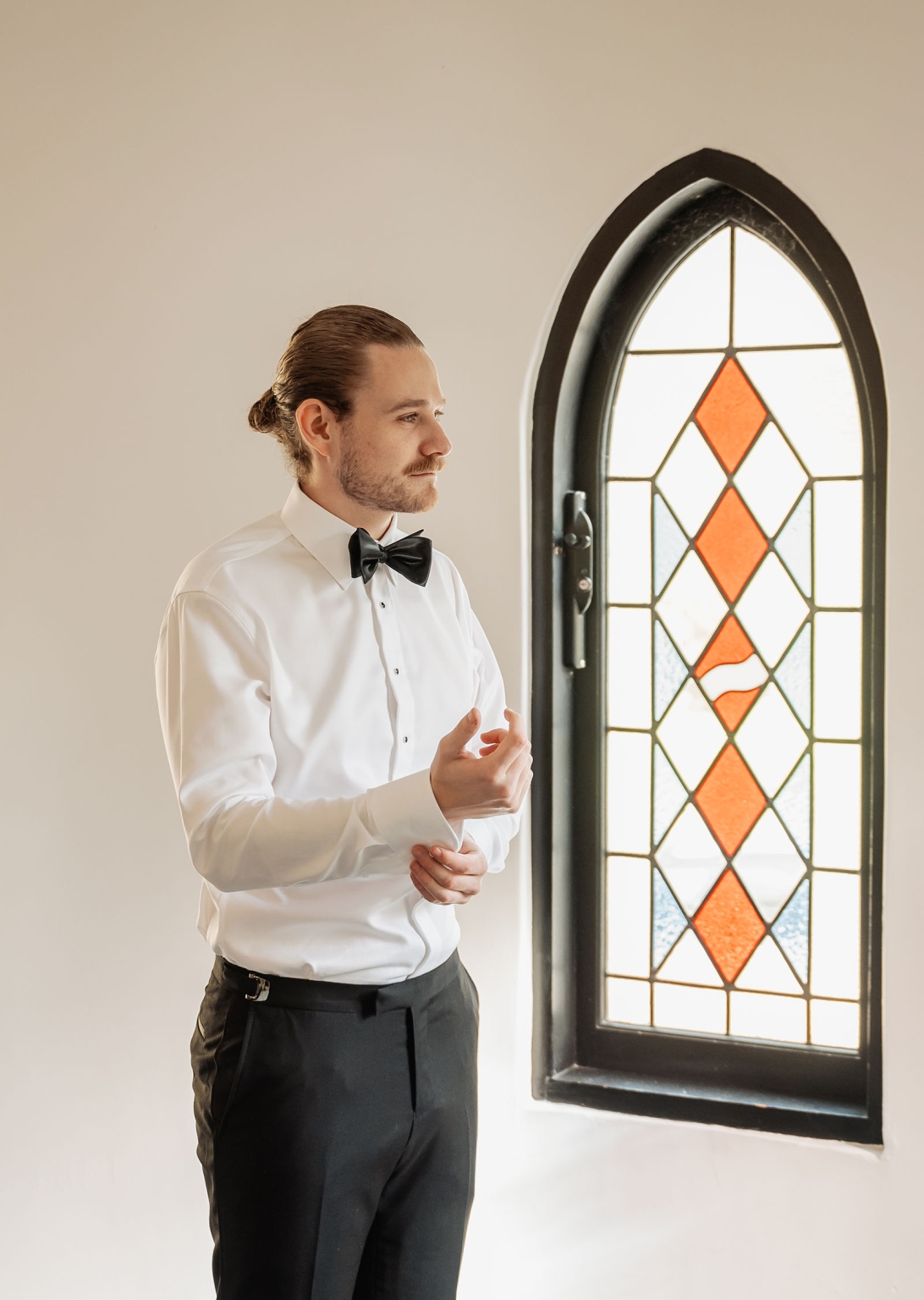 the manor wedding - crystal & mackenzie 5 Groom adjusting shirt cuff beside arched stained glass window in formal wedding attire.