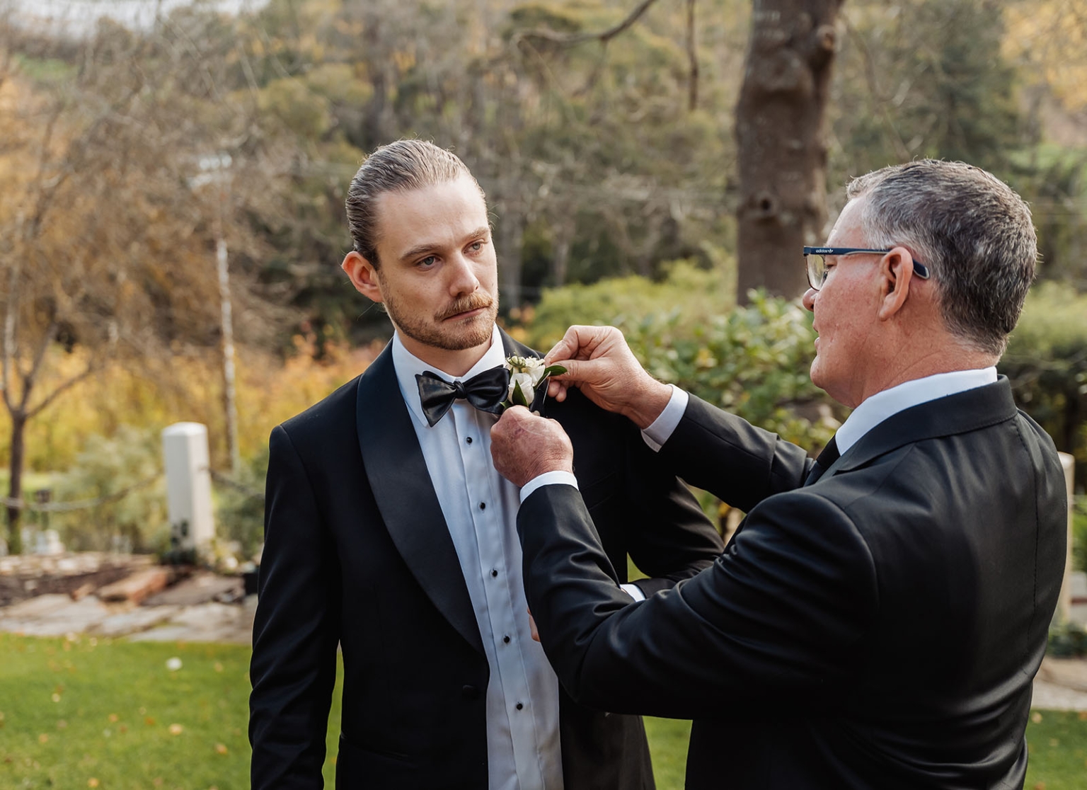 the manor wedding - crystal & mackenzie 9 Older man adjusts groom’s white boutonniere on black tuxedo at outdoor wedding.