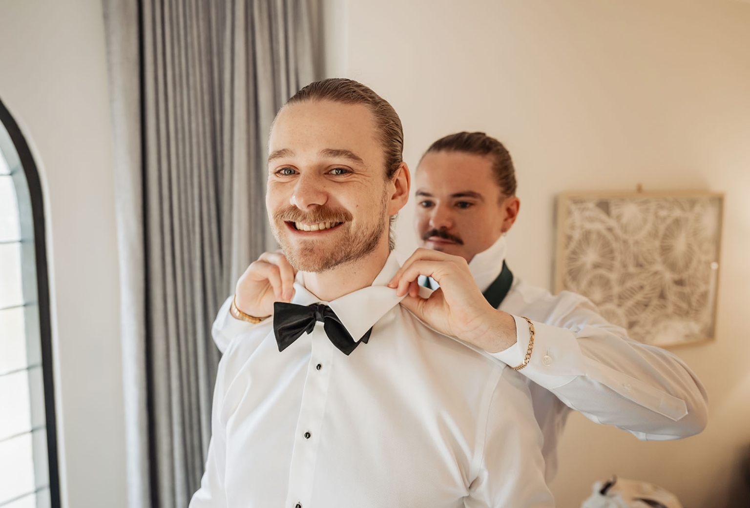 the manor wedding - crystal & mackenzie 3 Groom smiling as groomsman adjusts black bow tie before wedding.