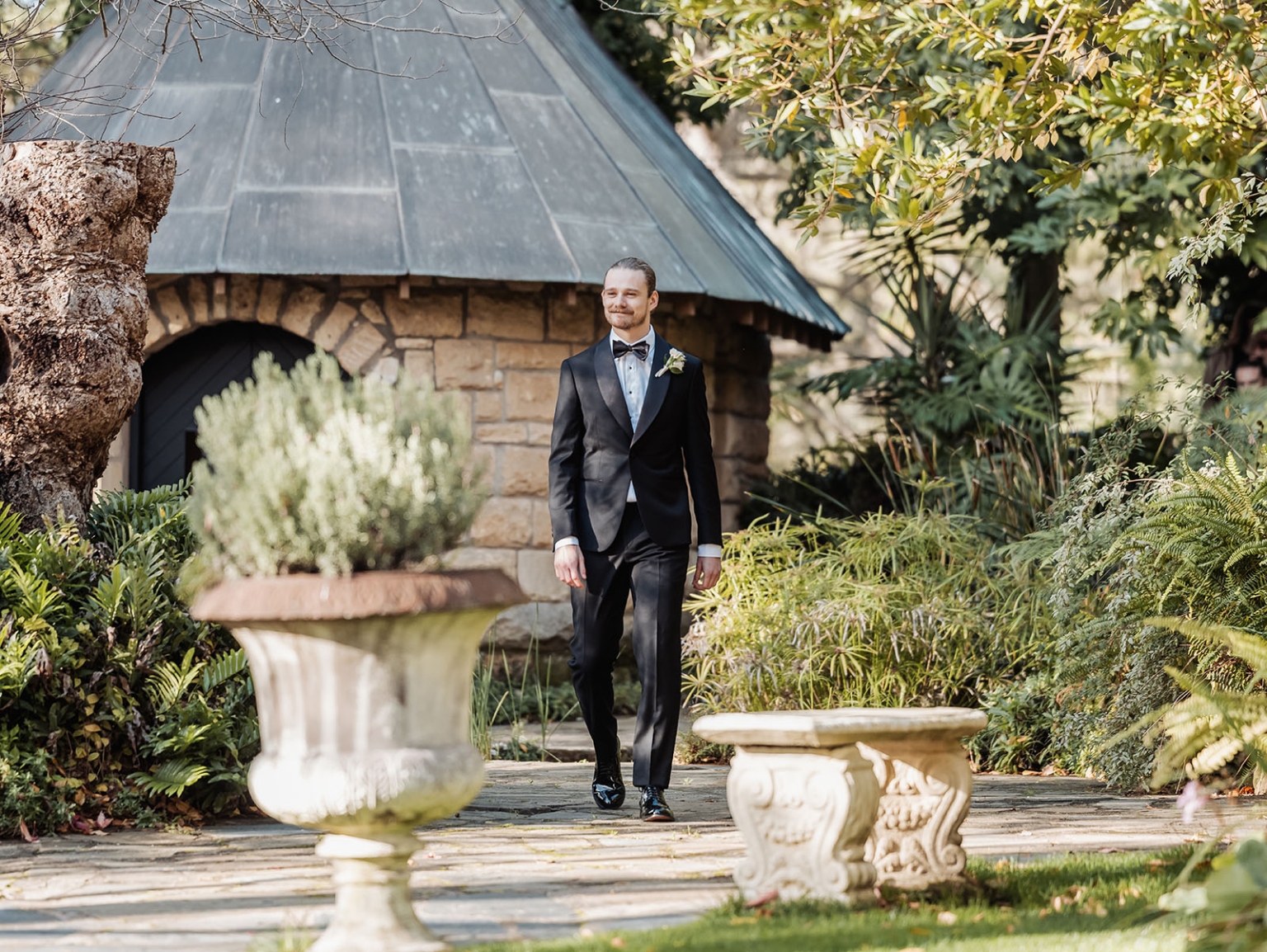 the manor wedding - crystal & mackenzie 46 Groom in black tuxedo walking through sunlit garden wedding venue.