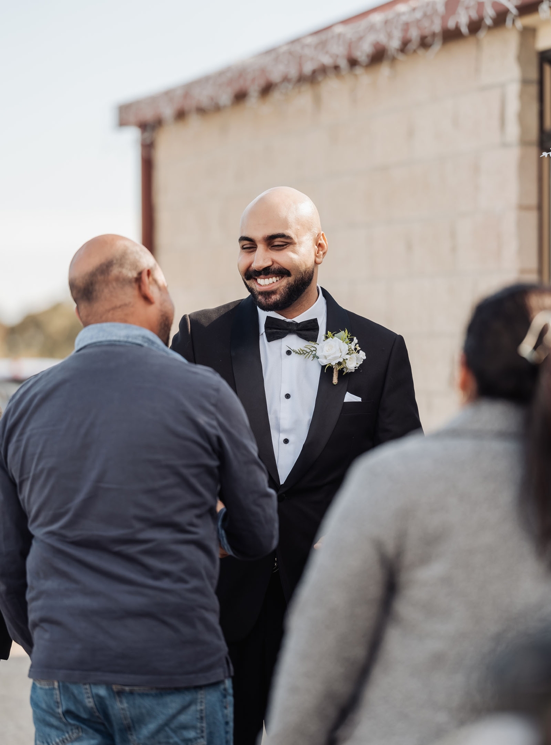 st george & st shenouda coptic church wedding - carmina & bill 32 Smiling groom in black tuxedo greets wedding guest at outdoor celebration.