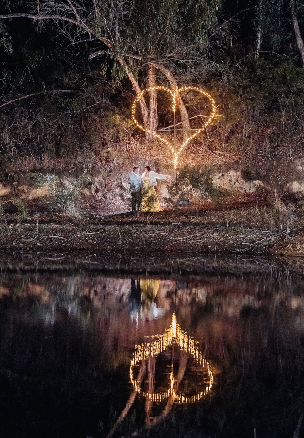 tiny house on the prairie wedding - lauren & bek 52 Romantic couple under heart-shaped string lights by reflective water at night.