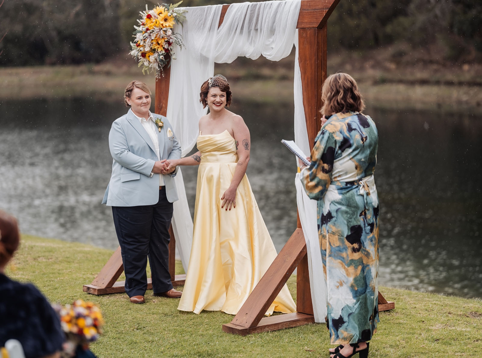 tiny house on the prairie wedding - lauren & bek 25 Lakeside wedding ceremony under floral wooden arch with couple and officiant.