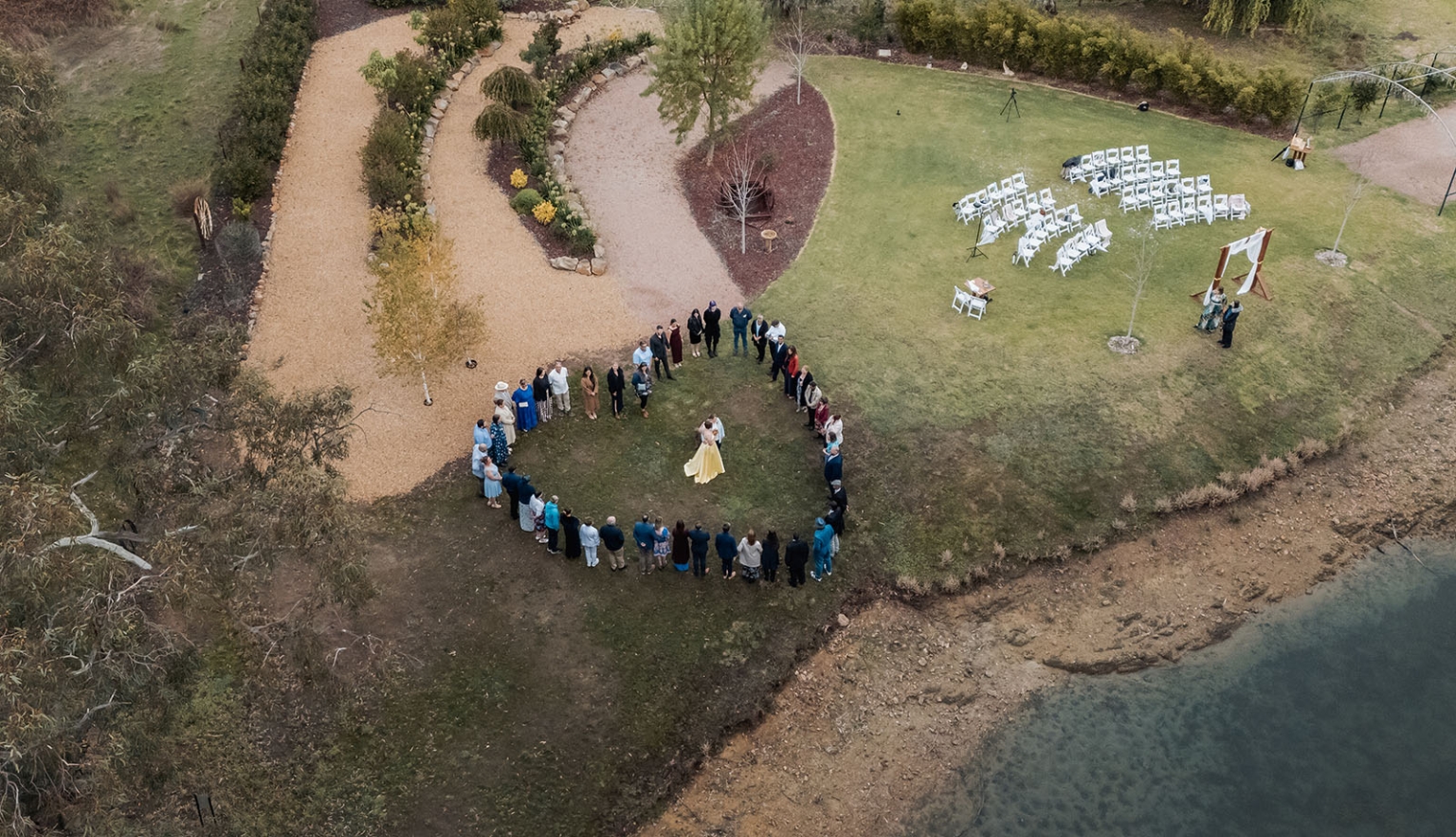tiny house on the prairie wedding - lauren & bek 37 Aerial outdoor lakeside wedding ceremony with guests in circle and white chairs.