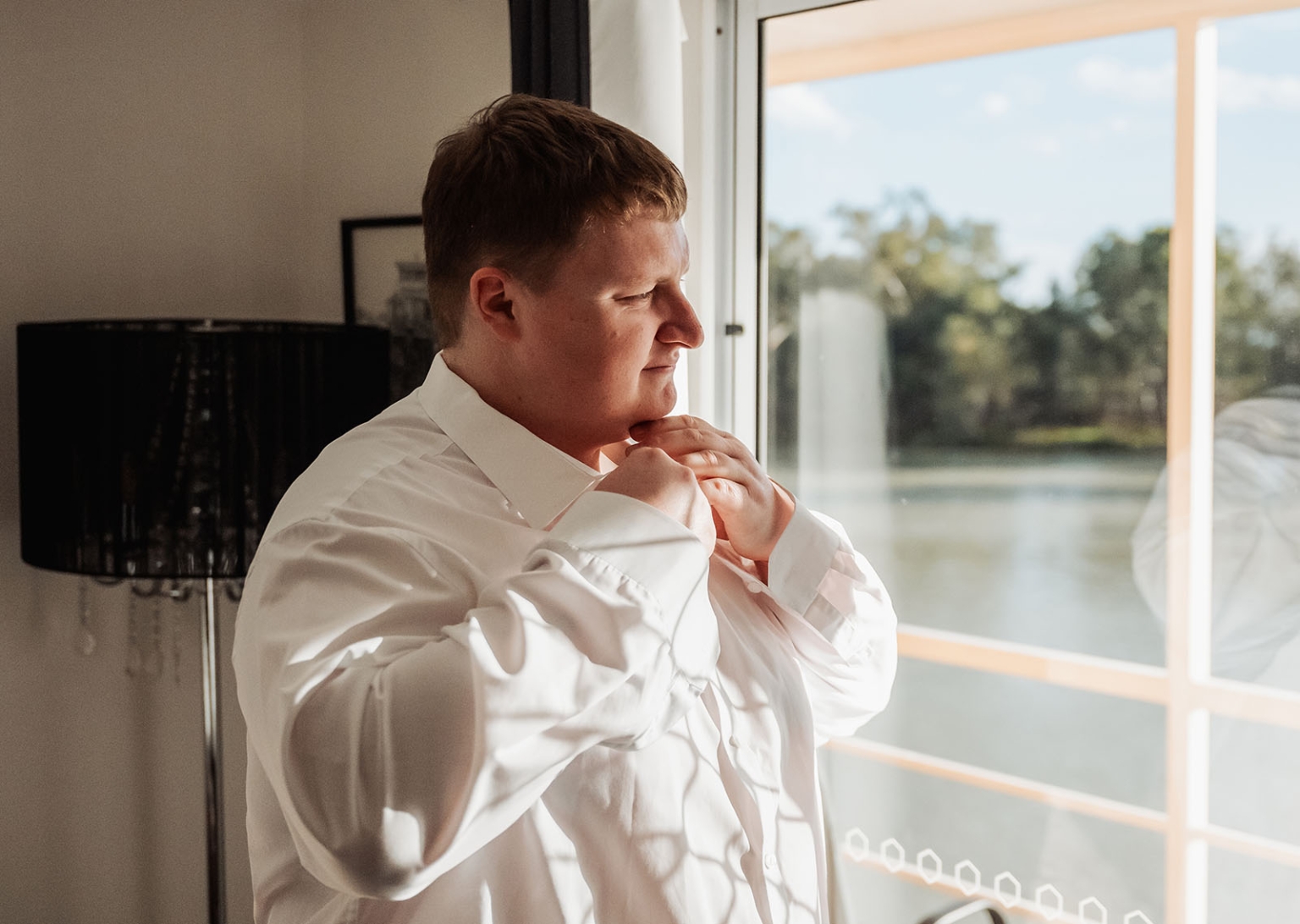loxton's st peter's church wedding - madeline & brayden 3 Groom adjusting white dress shirt by sunlit window before wedding.