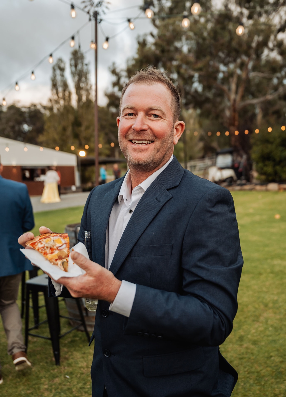 tiny house on the prairie wedding - lauren & bek 57 Man in suit eating pizza at outdoor wedding reception with string lights.