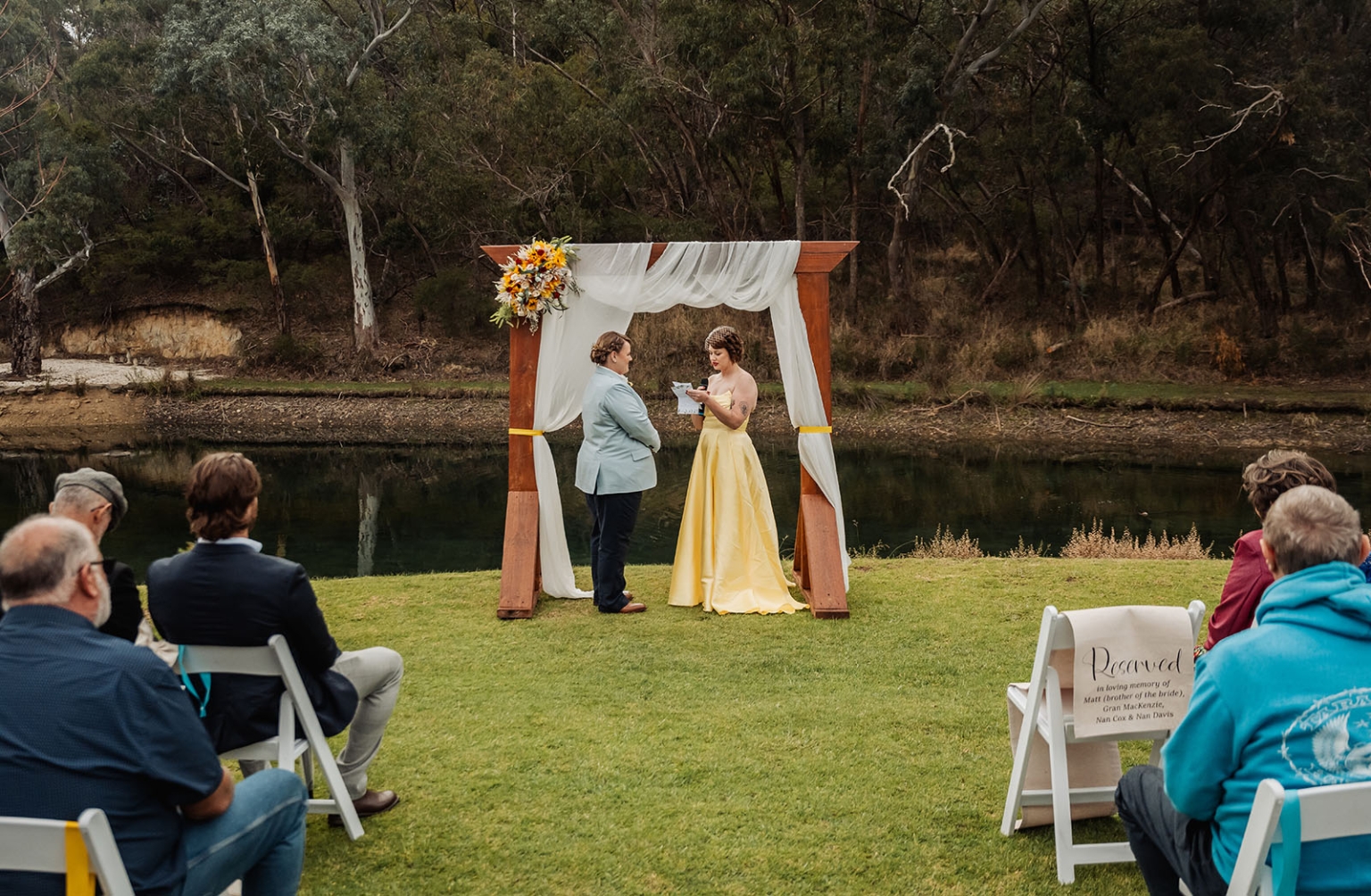 tiny house on the prairie wedding - lauren & bek 31 Outdoor riverside wedding ceremony with wooden arch, yellow dress, and seated guests.