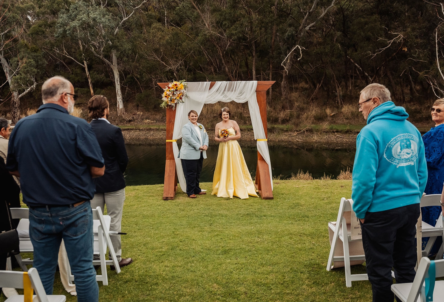 tiny house on the prairie wedding - lauren & bek 22 Outdoor riverside wedding ceremony with floral arch, yellow bridal gown, and guests.