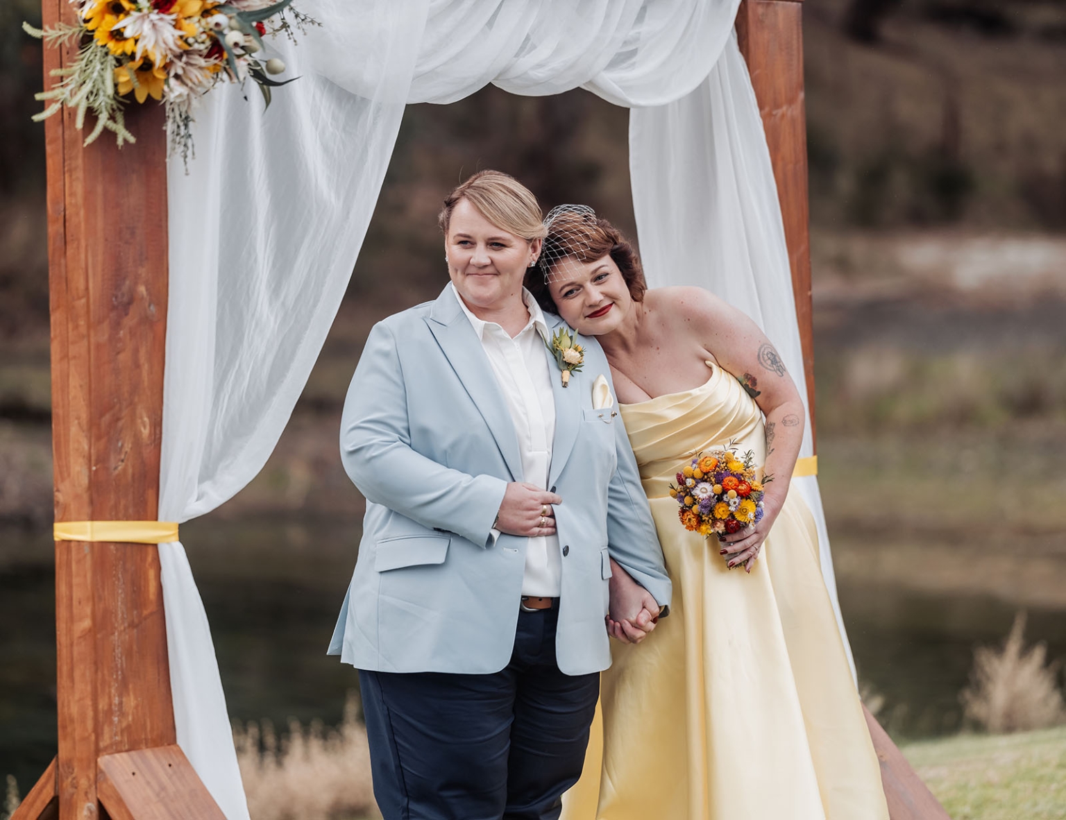 tiny house on the prairie wedding - lauren & bek 23 Newlyweds pose under rustic outdoor wedding arch with sunflowers and bouquet.