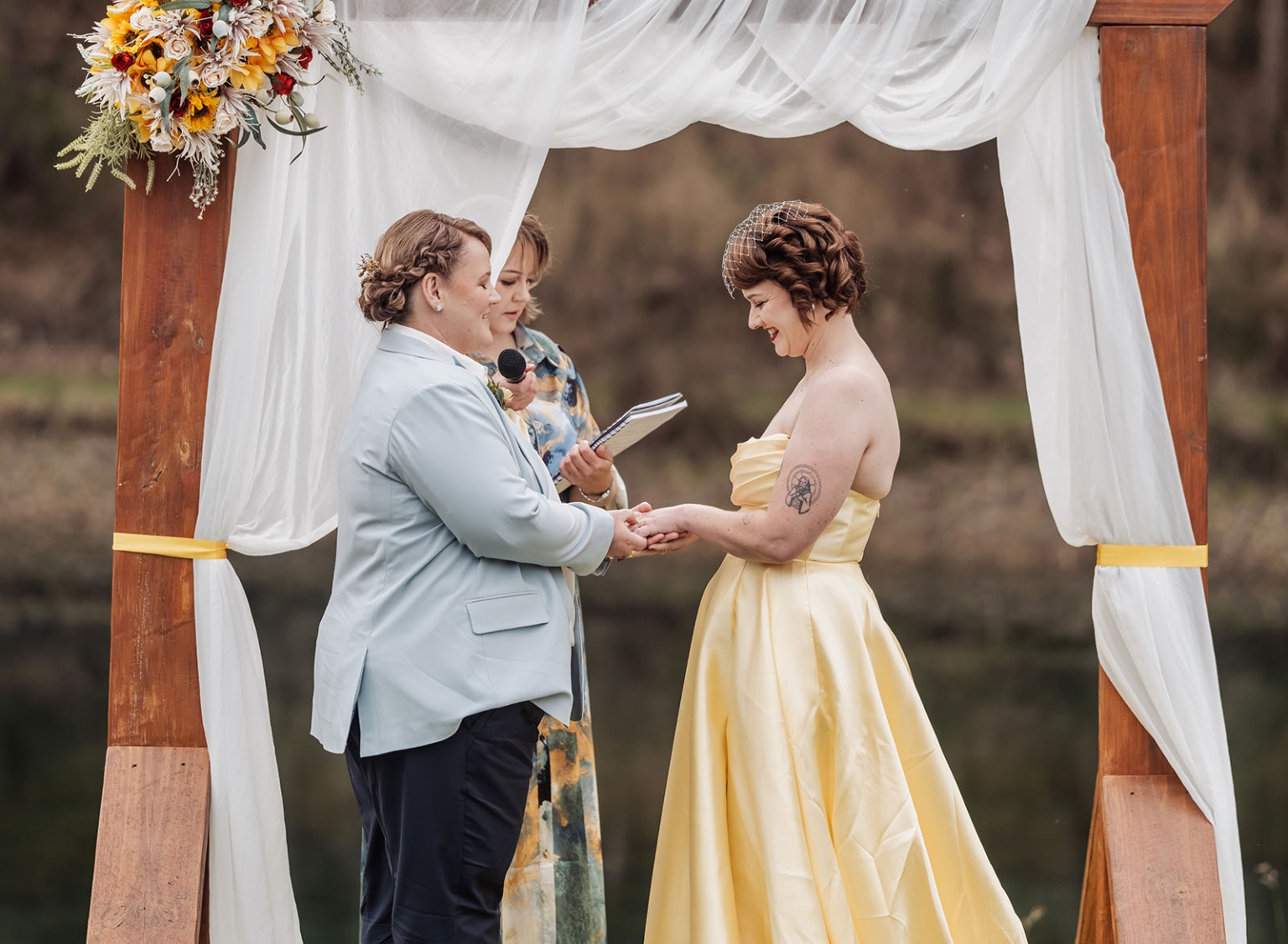 tiny house on the prairie wedding - lauren & bek 33 Two brides exchange vows under floral wooden arch at outdoor wedding ceremony.