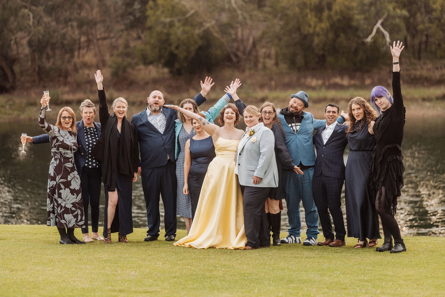 tiny house on the prairie wedding - lauren & bek 54 Joyful outdoor wedding group photo by a lake with cheering guests.