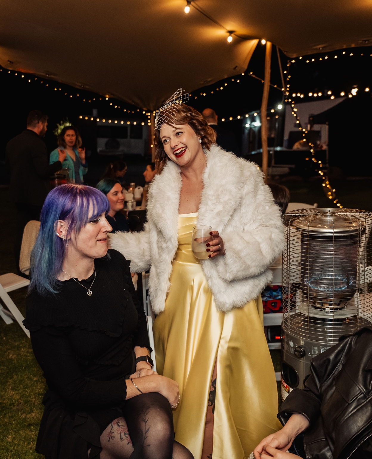 tiny house on the prairie wedding - lauren & bek 61 Outdoor wedding reception under string lights with bride laughing beside guests and patio heater.