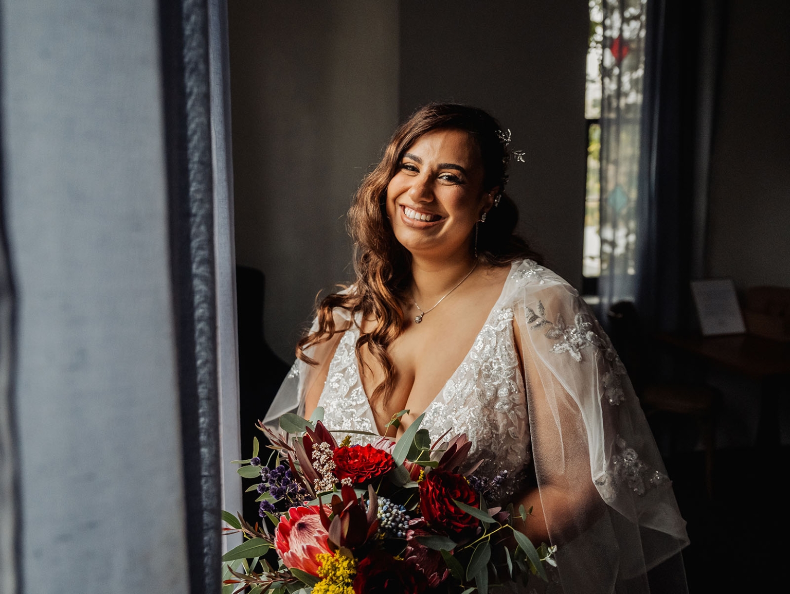 basket range wedding at the manor - leah & wes 11 Smiling bride in lace wedding gown holding burgundy bouquet by sunlit window.