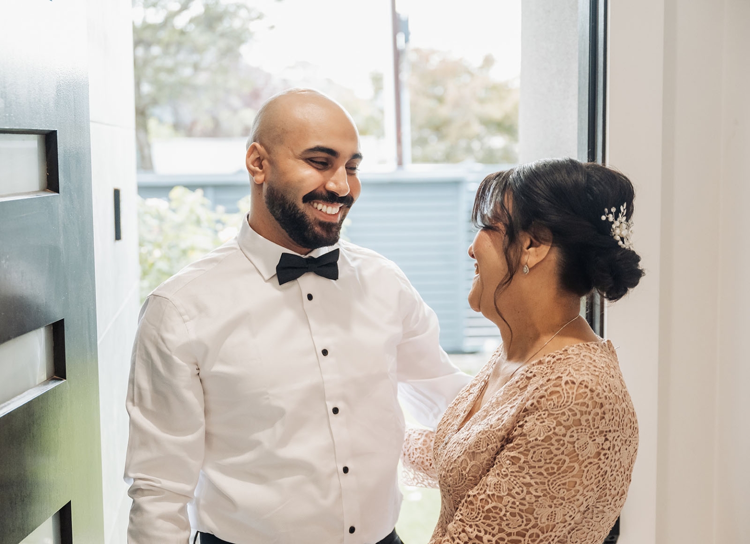 st george & st shenouda coptic church wedding - carmina & bill 6 Joyful wedding couple in formal attire sharing a romantic moment by a sunlit window.